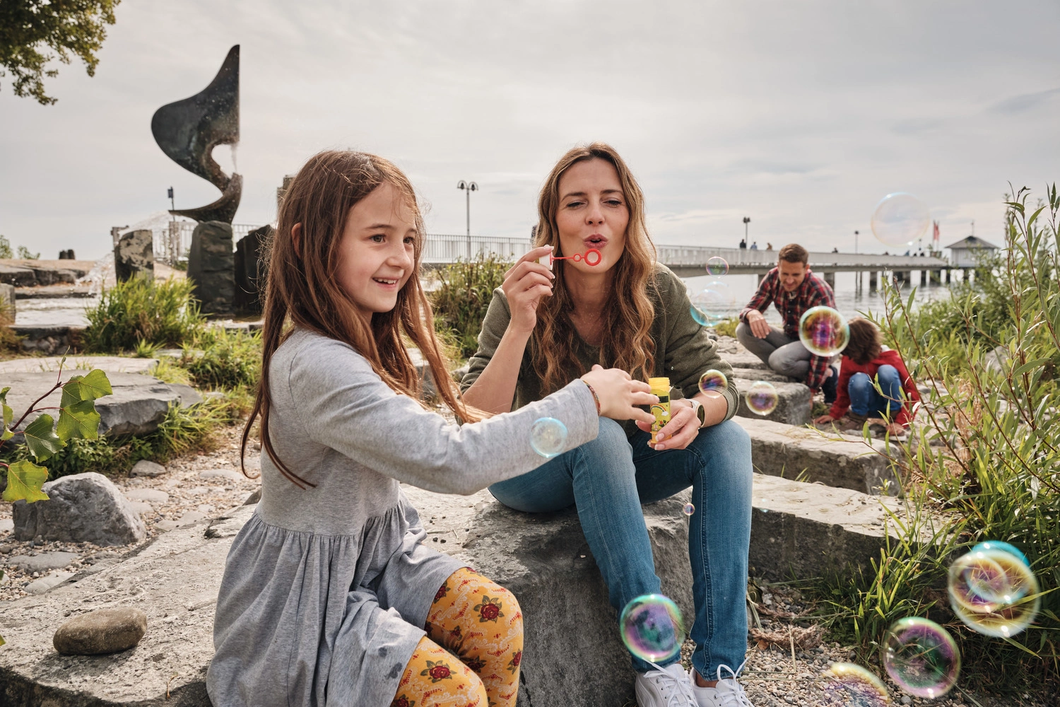 Mutter und Tochter sitzen am Ufer eines Sees und machen Seifenblasen. Im Hintergrund ein Landungssteg.