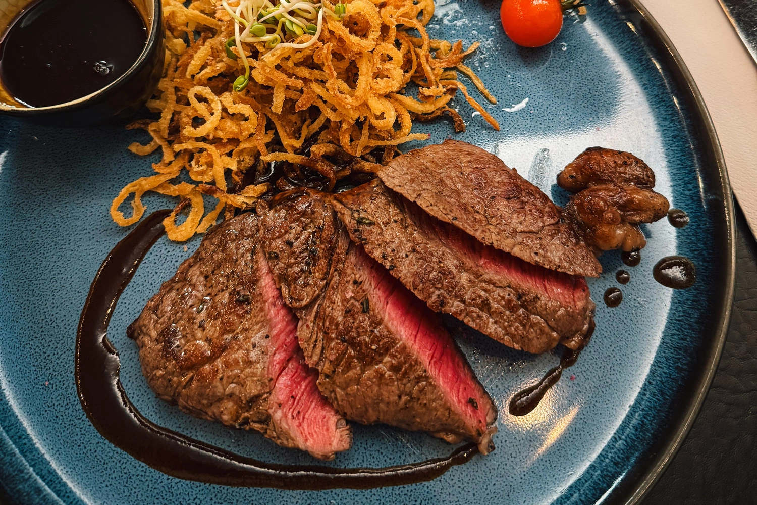 Sliced, pink roasted beef steak with dark sauce, crispy fried onions, mushrooms, and a cocktail tomato on a blue plate—served as a fine main course.