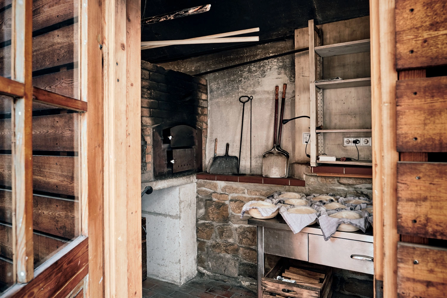 Blick durch eine Tür in den Innenraum eines alten Backhauses. Zu sehen ist ein alter Steinofen, Feuerholz und auf einer Ablage liegen Brote in Formen und warten darauf in den ofen geschoben zu werden.