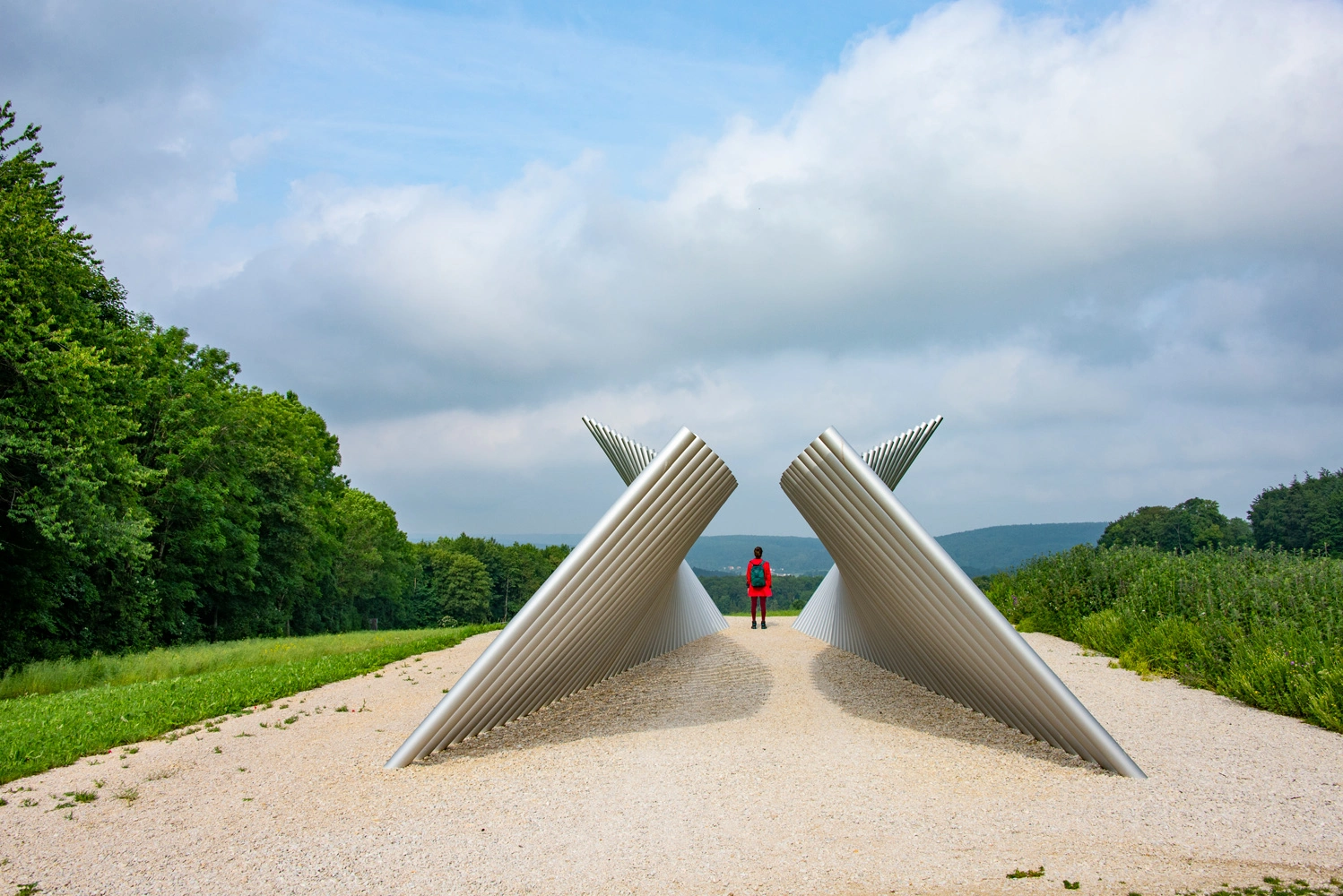 A person in a red jacket is standing between two sculptures. The sculptures are made of different metal tubes arranged diagonally in a row.