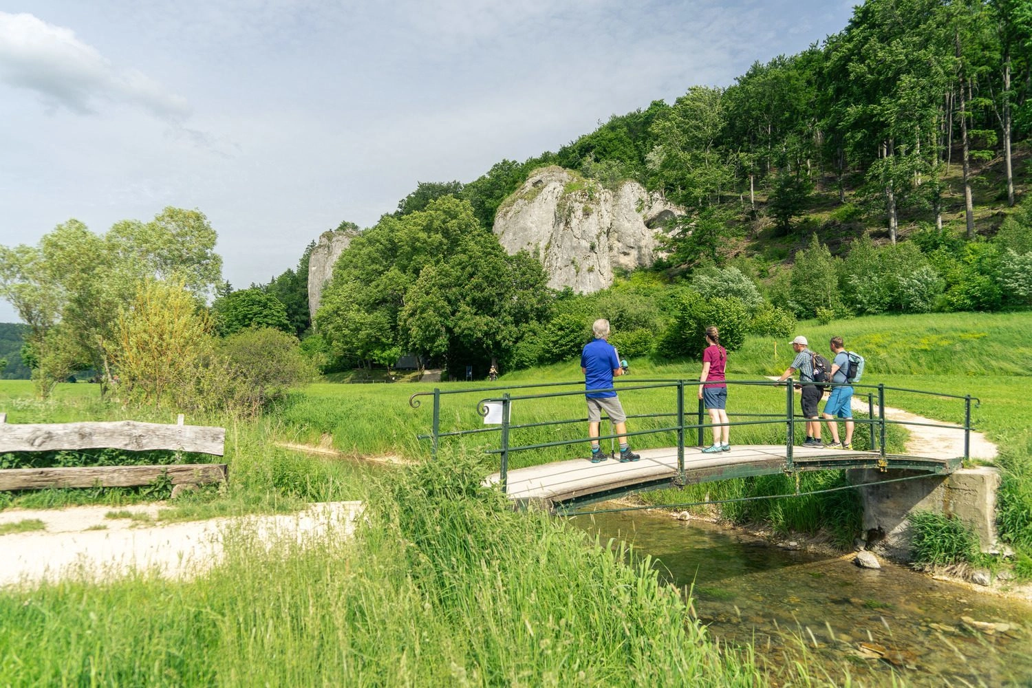 Wanderer stehen auf einer Brücke über einen kleinen Fluss und blicken zu einer Felsformation.
