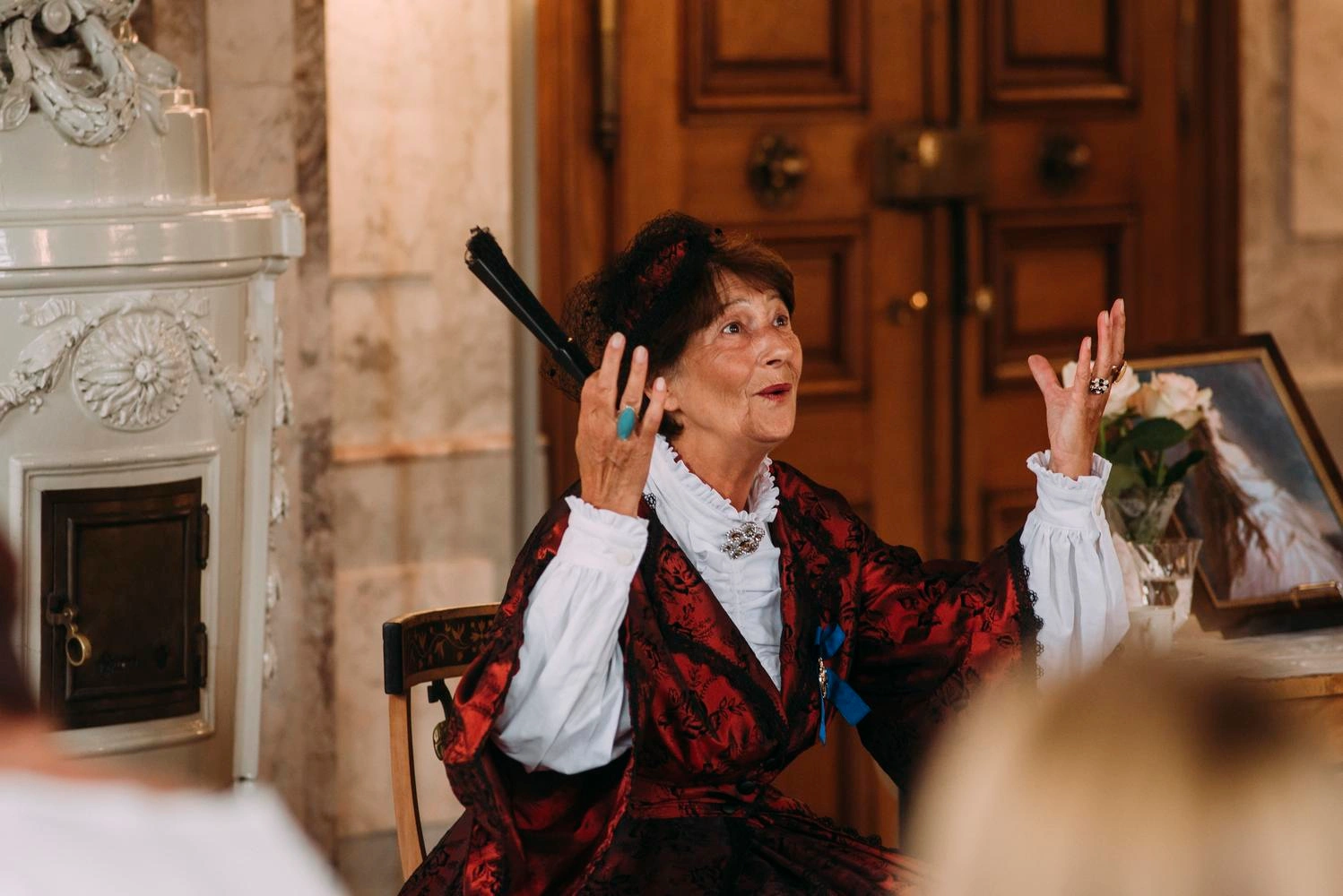 An elderly lady is sitting in a room of a castle with a fan in her hand and her arms raised.