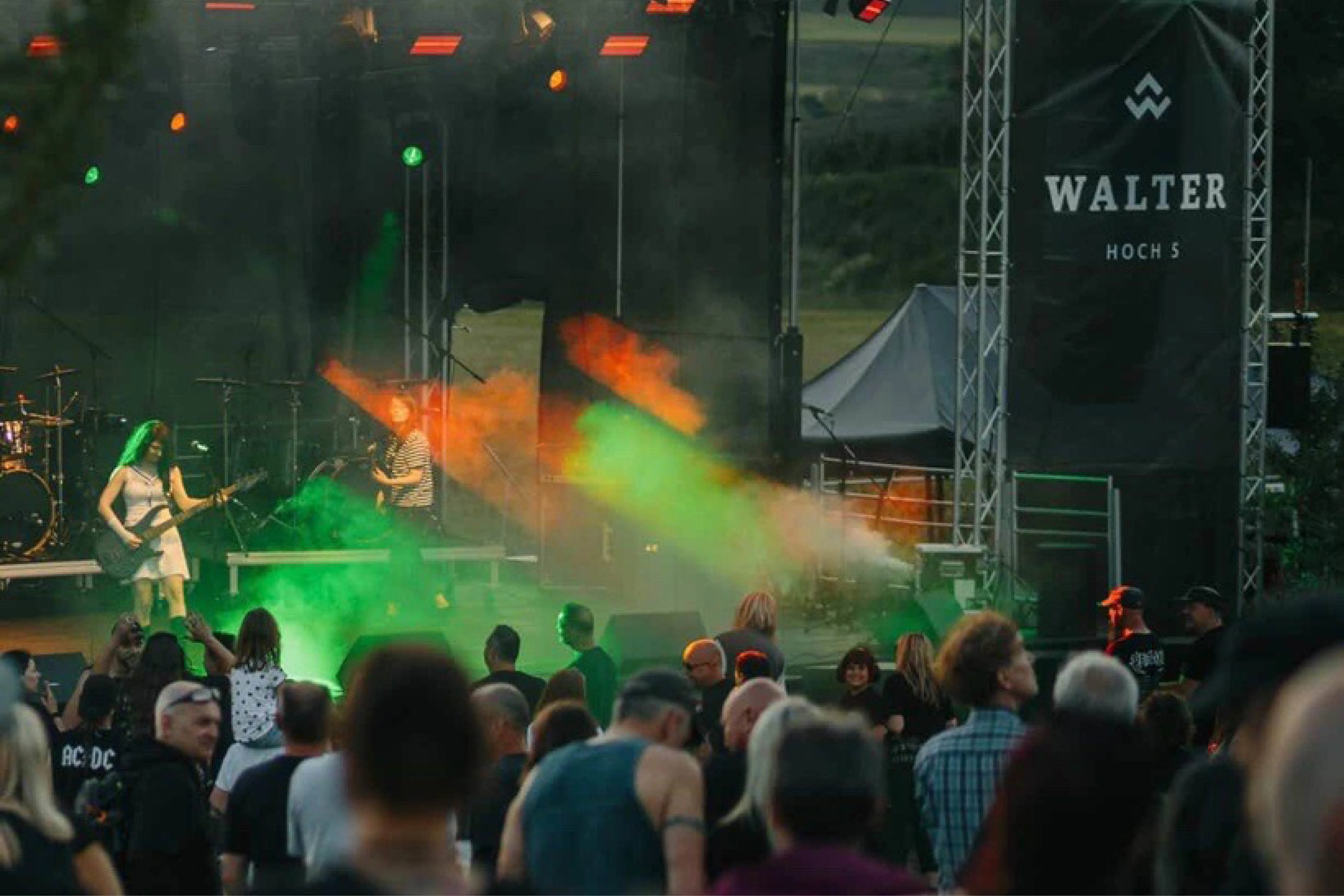 Open-Air-Konzert mit Gitarristin und Band auf Bühne, farbige Licht- und Nebeleffekte; Publikum steht davor, Banner „WALTER HOCH 5“ im Hintergrund.