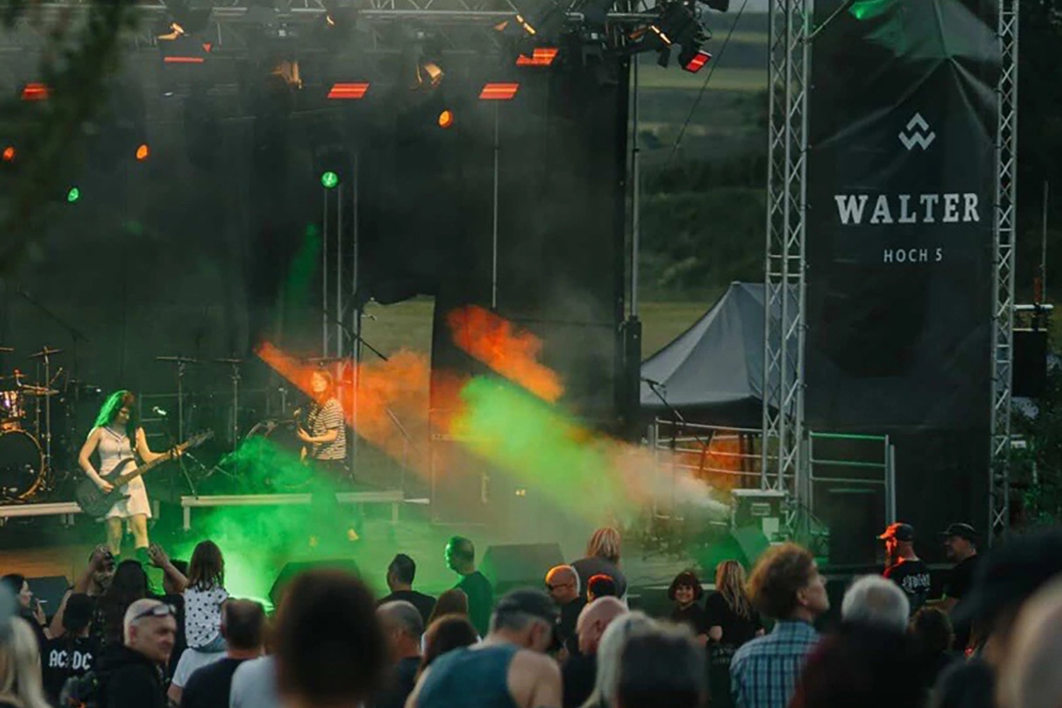 Open-Air-Konzert mit Gitarristin und Band auf Bühne, farbige Licht- und Nebeleffekte; Publikum steht davor, Banner „WALTER HOCH 5“ im Hintergrund.