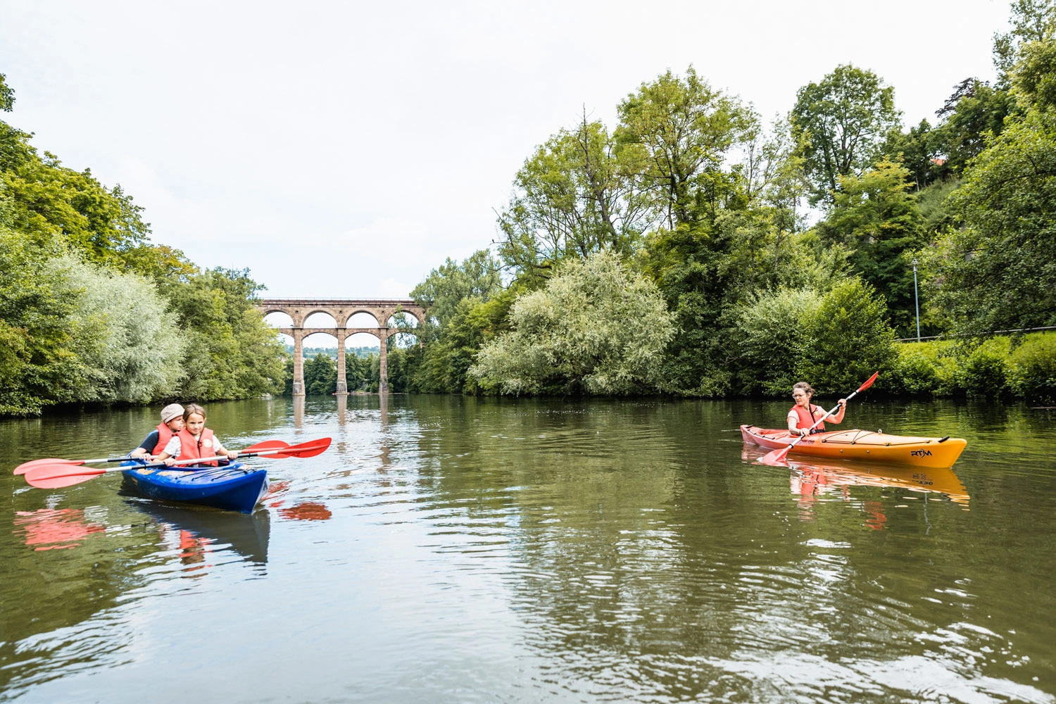 Zwei Kinder und eine Mutter paddeln in Kanus auf einem Fluss. Im Hintergrund sieht man ein Viadukt.