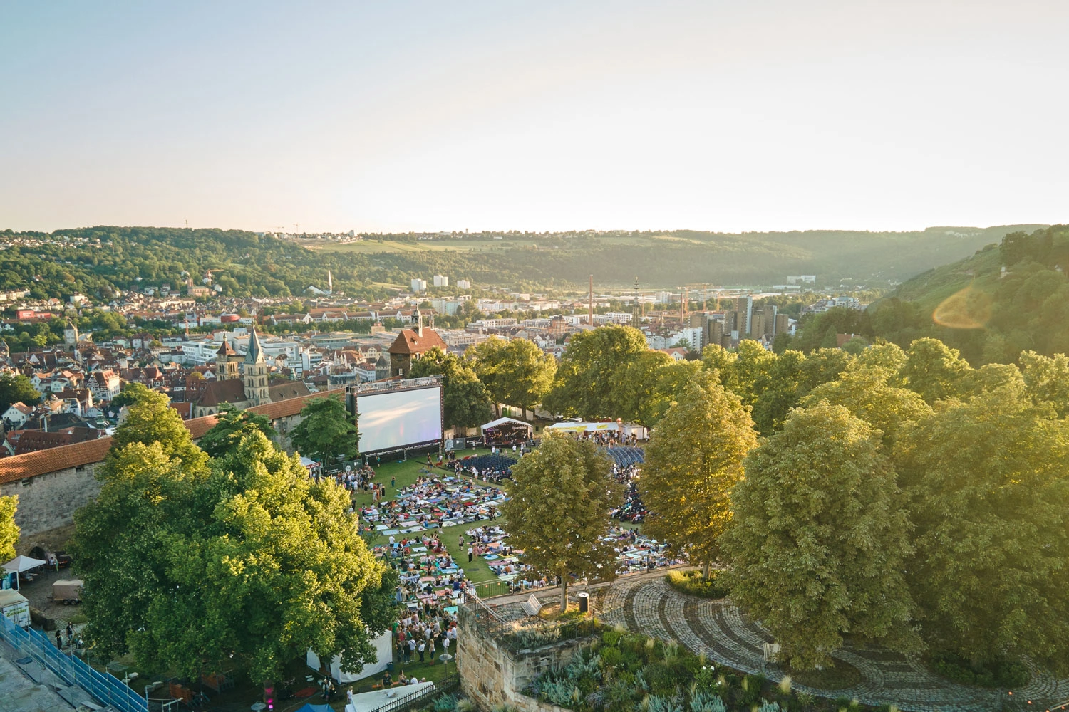 A panoramic view of a city with an open-air movie theater on a hill: a large screen, numerous visitors on a lawn, and a sunny hilly landscape in the background.