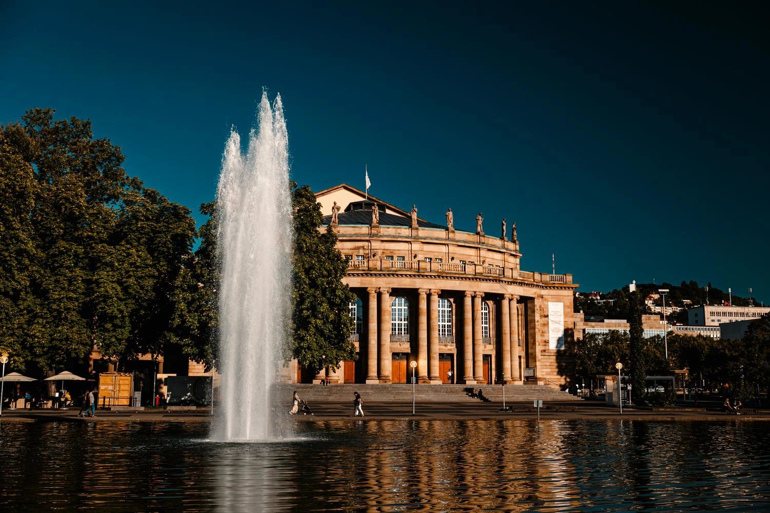 Historisches Opernhaus mit klassischer Fassade und Säulen am Eckensee in Stuttgart; im Vordergrund ein hoher Springbrunnen, umgeben von Bäumen und spiegelndem Wasser bei klarem Himmel.