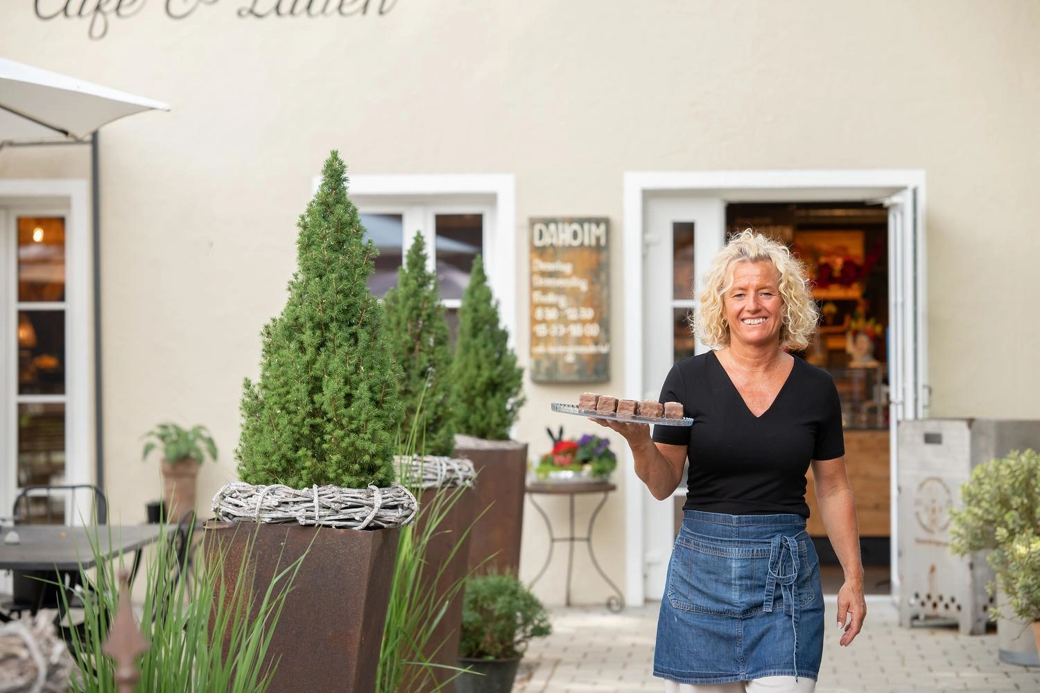 Person trägt ein Tablett mit Gebäck vor dem Eingang eines Hofcafés mit Pflanzen und geöffneter Tür.