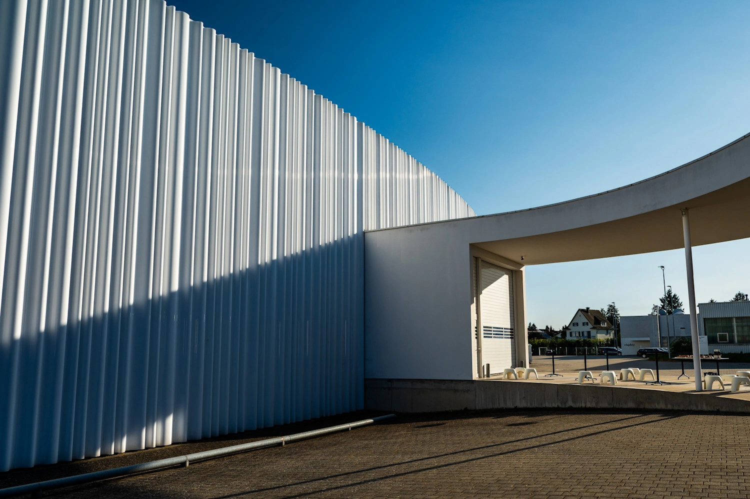 Der überdachte Eingang zu einer großen, weißen Halle, dessen Fassade gewellt ist. 