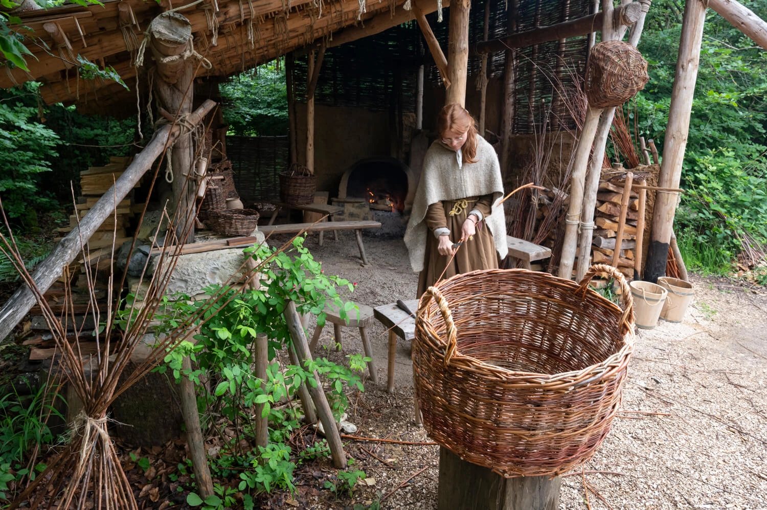 A woman in medieval clothing stands under a wooden shelter. She is holding a thin branch with which she is weaving baskets. In front of the woman is a large woven basket.