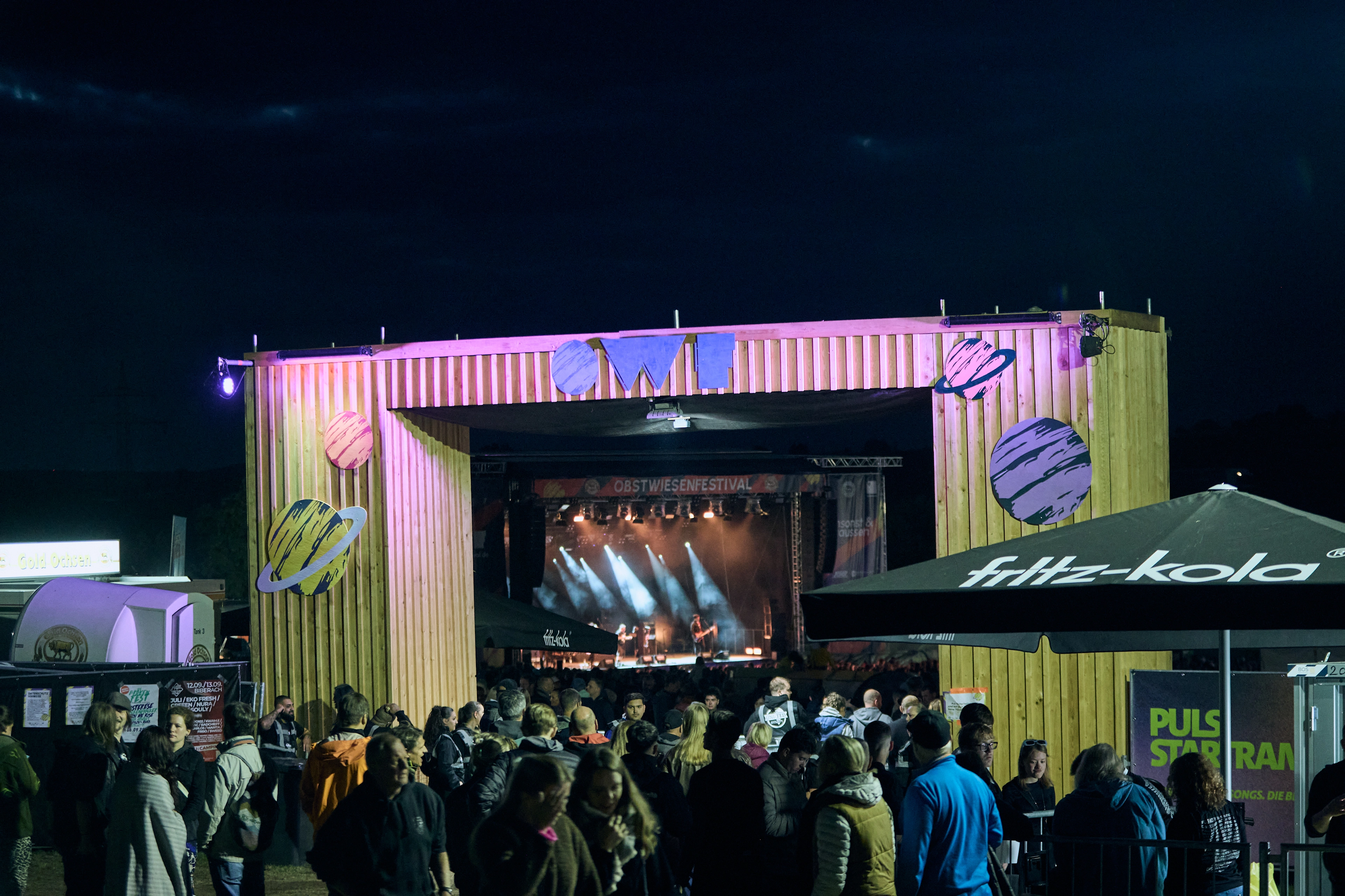 Eingangsbereich des Obstwiesenfestivals in Dornstadt bei Ulm mit Holzportal „OWF“, Besucherströmen und beleuchteter Konzertbühne im Hintergrund.