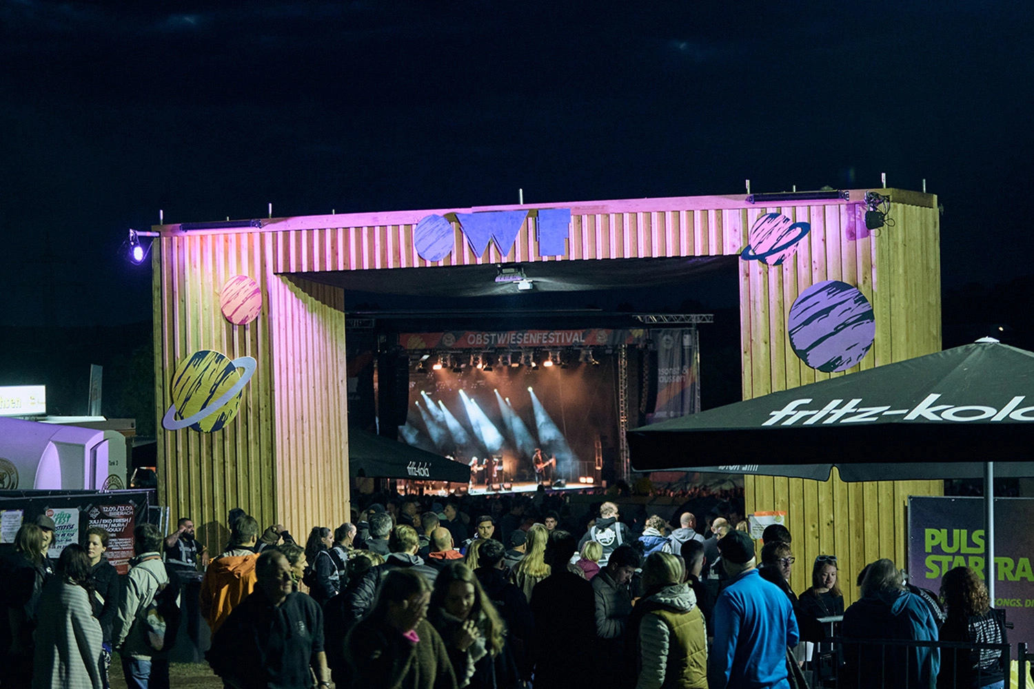 Eingangsbereich des Obstwiesenfestivals in Dornstadt bei Ulm mit Holzportal „OWF“, Besucherströmen und beleuchteter Konzertbühne im Hintergrund.