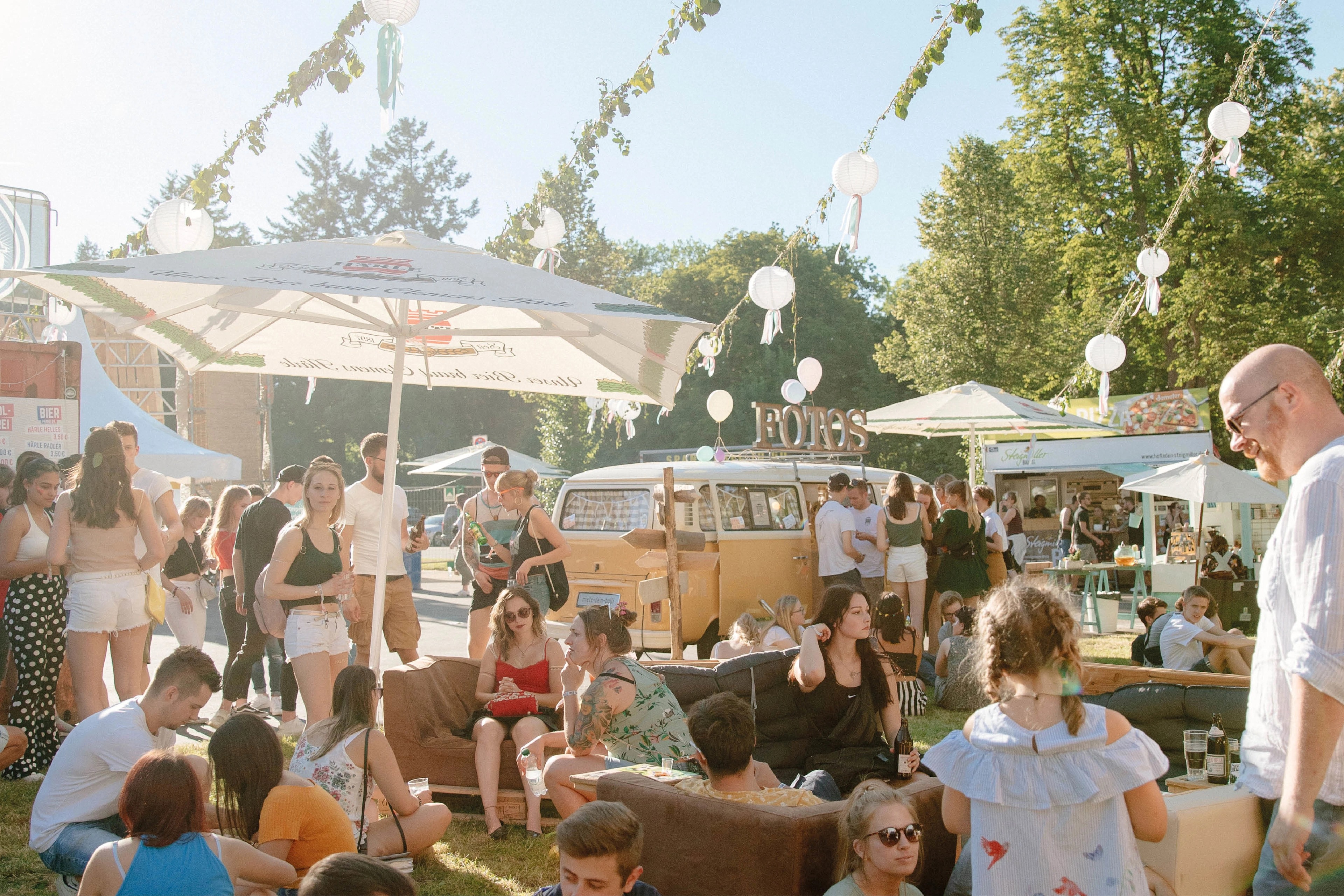 Festivalbesucher entspannen im Außenbereich mit Foodtrucks und Foto-Bus; Lichterketten und Sonnenschirme schaffen sommerliche Atmosphäre.