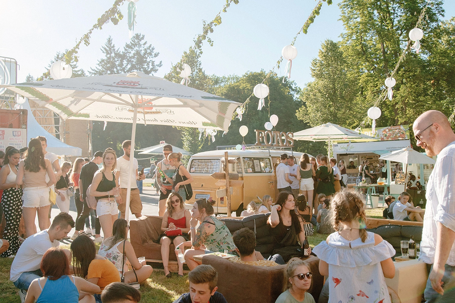 Festivalbesucher entspannen im Außenbereich mit Foodtrucks und Foto-Bus; Lichterketten und Sonnenschirme schaffen sommerliche Atmosphäre.