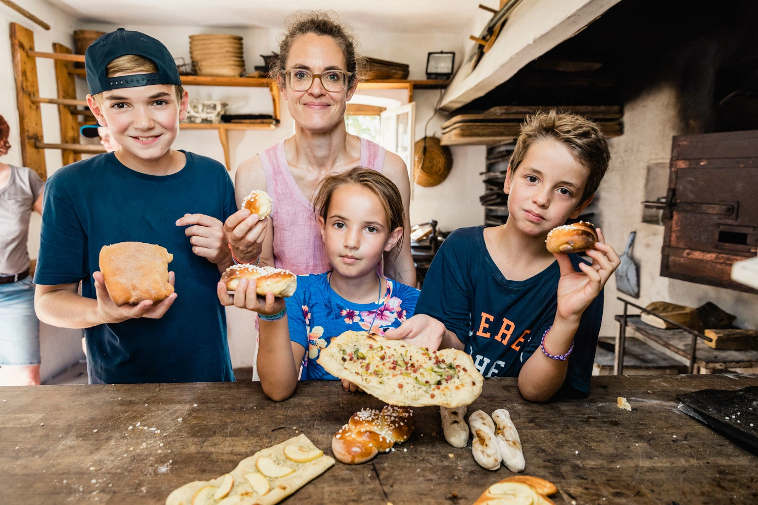 Eine Frau steht mit ihren drei Kindern hinter einem großen Tisch in einer alten Backstube. Alle halten ihre selbstgemachten Gebäckteile in die Höhe.