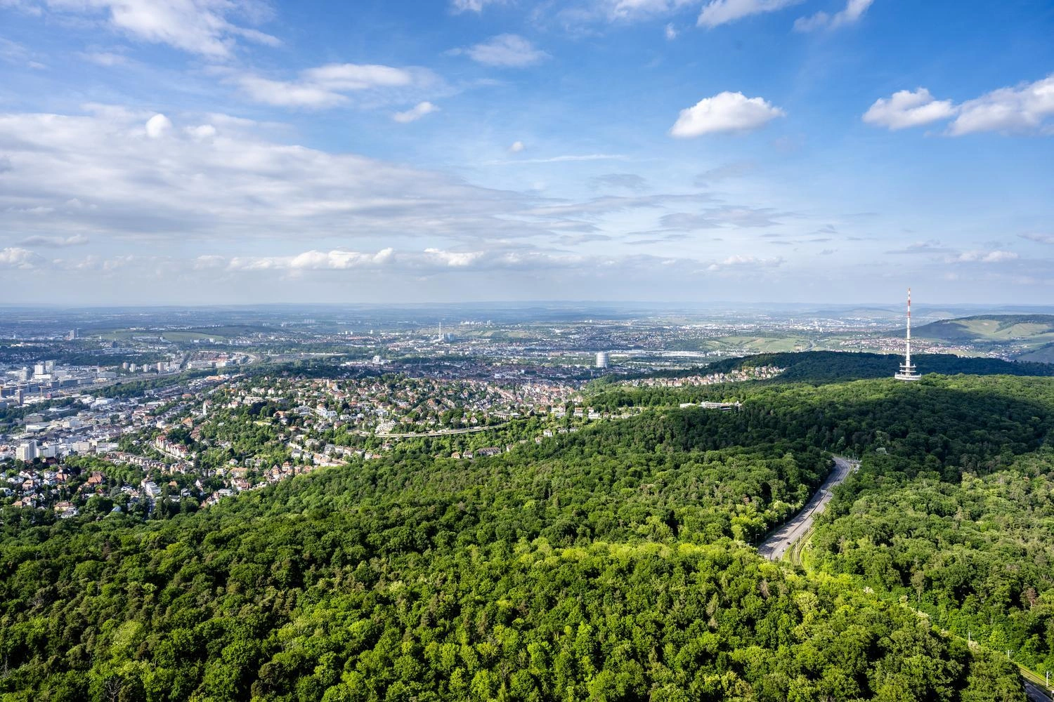 Blick über Stuttgart mit dichtem Wald im Vordergrund, Stadt im Hintergrund und Turm unter blauem Himmel mit Wolken.