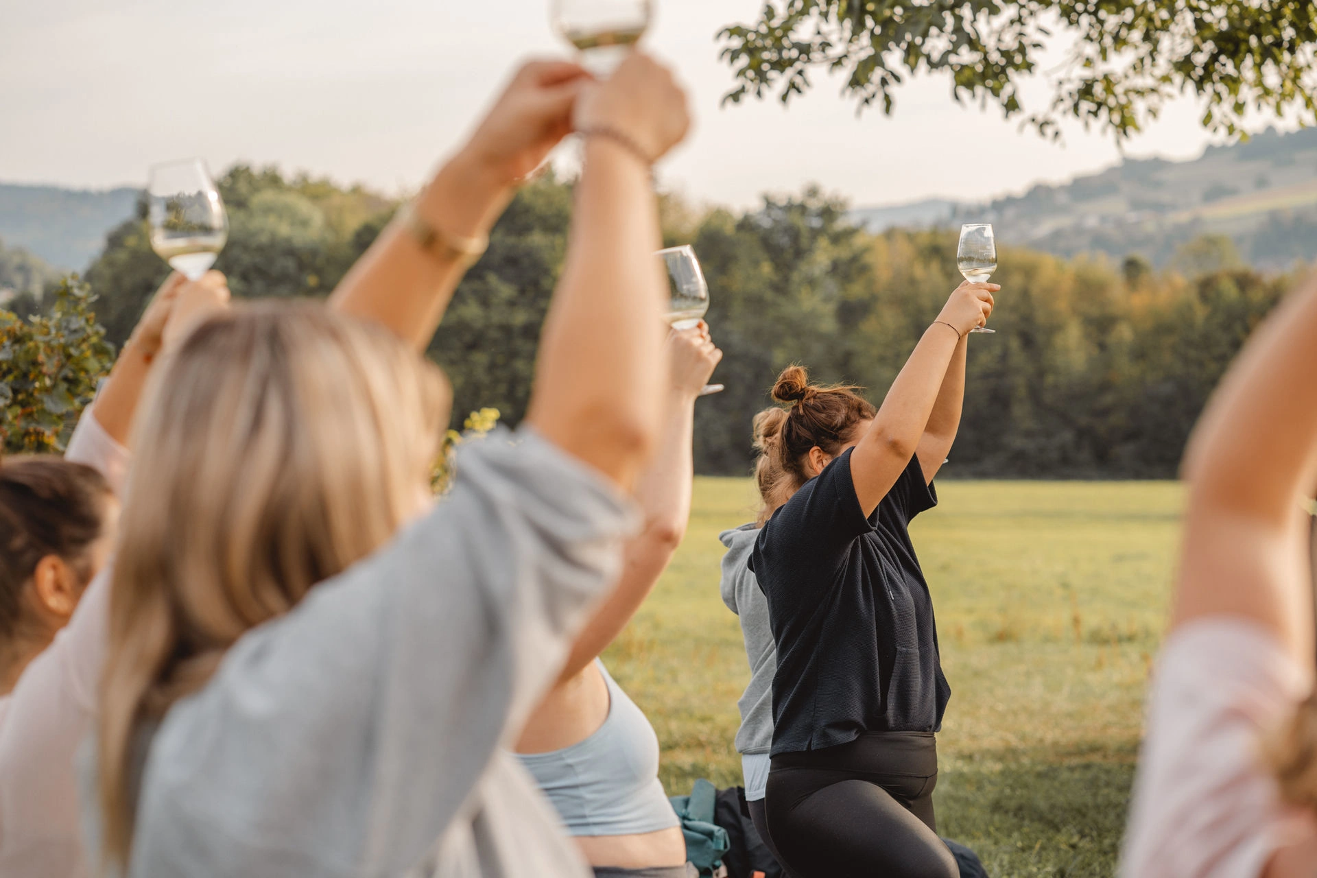 Mehrere Personen im Freien verharren in einer Yoga-Pose und halten dabei ein Weinglas über ihre Köpfe.