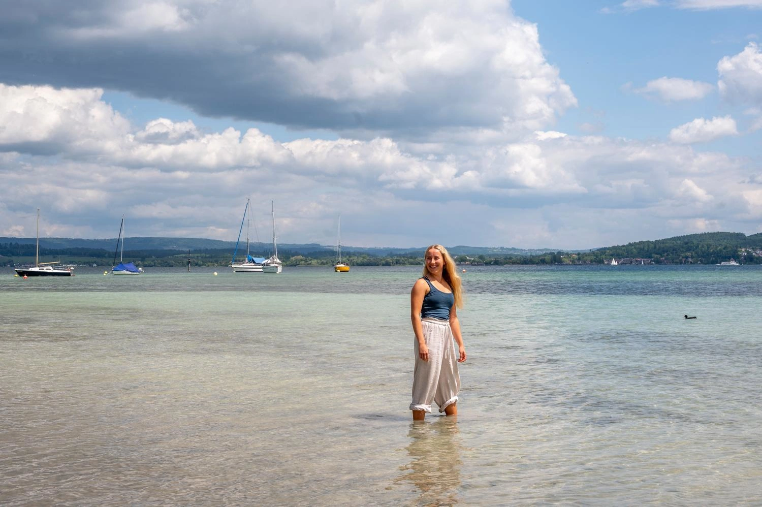 Person steht im flachen Wasser des Bodensees, umgeben von klarem Wasser, Segelbooten und bewaldeten Ufern unter teils bewölktem Himmel.