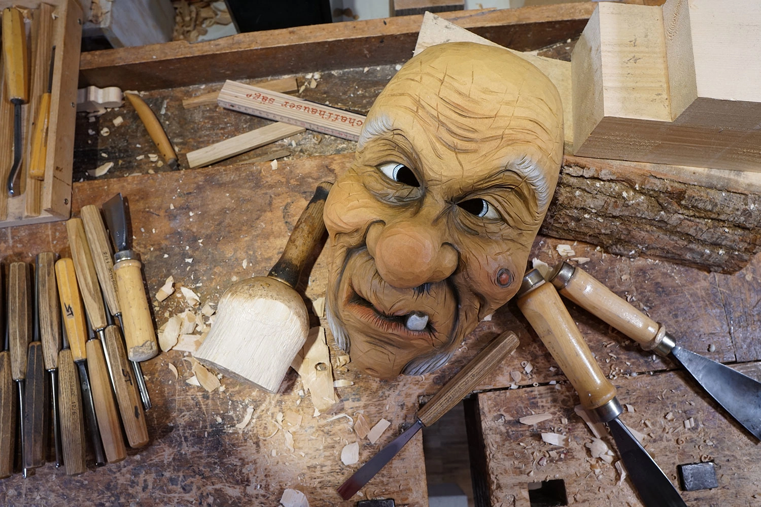 A finished carved wooden jester mask lies on the workbench. The tools used lie around it.