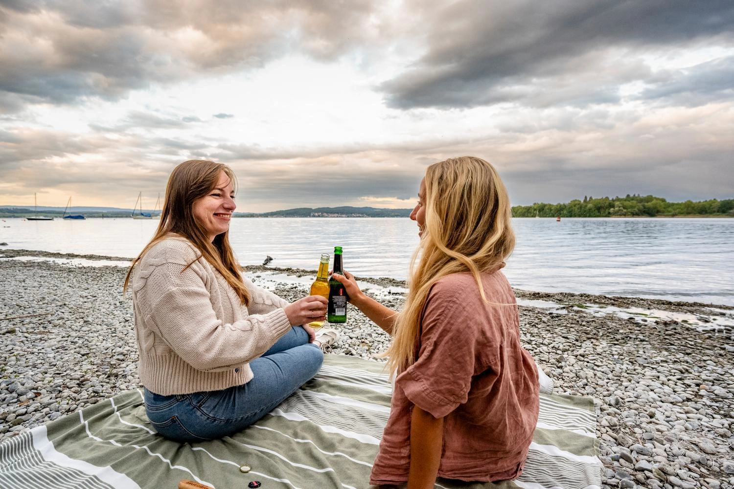 Zwei Personen sitzen auf einer Decke am Kiesstrand des Bodensees und stoßen mit Flaschen an, im Hintergrund ruhiges Wasser und bewaldetes Ufer unter wolkigem Himmel.
