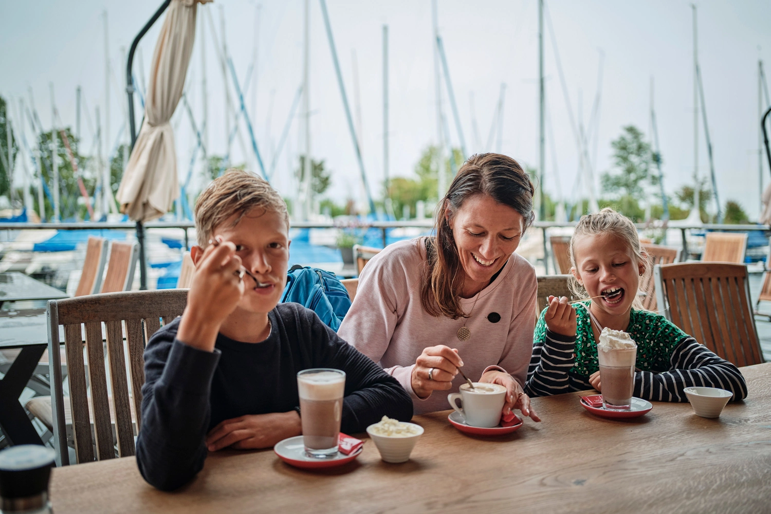 A mother is sitting with her two children on the terrace of a restaurant at a harbor. She is relaxing with a coffee and the children also have chocolate with cream in front of them.