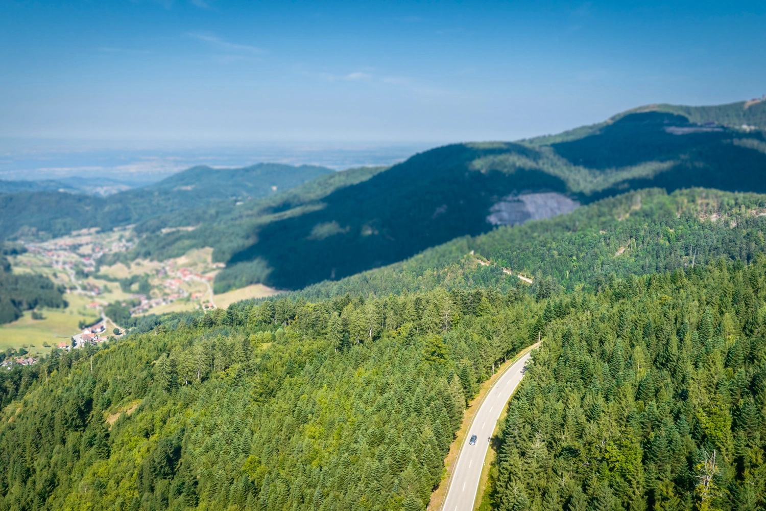 Aerial view of a winding road snaking through dense forest in the low mountain range, with sweeping views over wooded hills and valleys under blue skies.