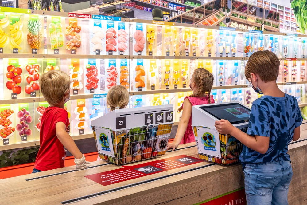 Children stand in front of a large wall on which different foods are displayed. In the foreground are two mesh baskets containing toy foodstuffs.