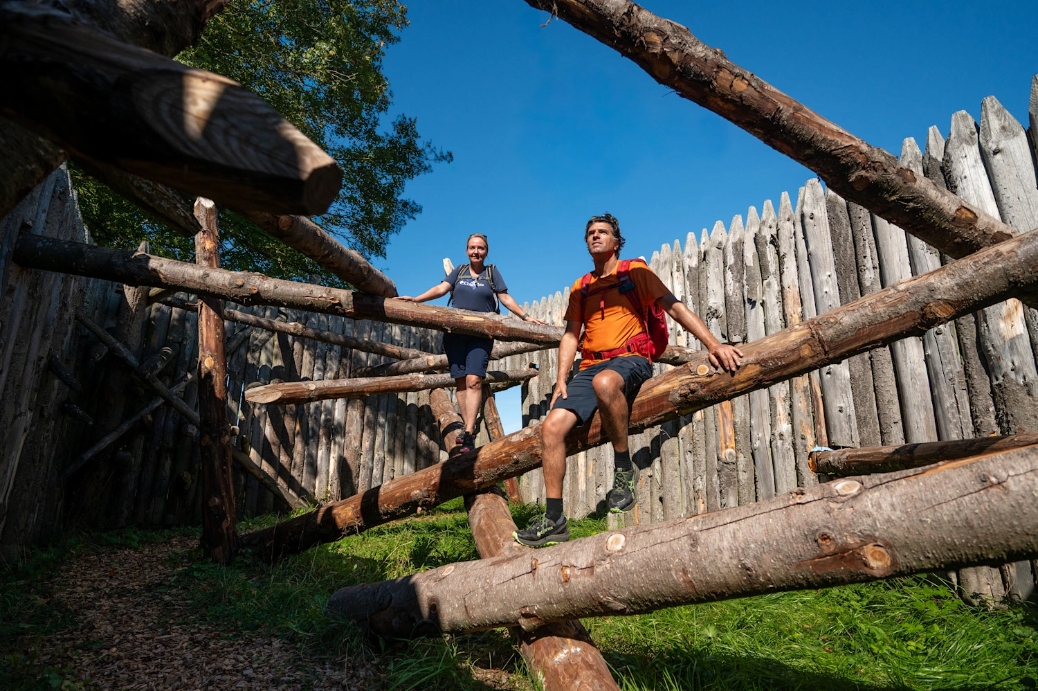 Zwei Personen in Wanderkleidung sitzen und stehen auf dicken Holzstämmen.