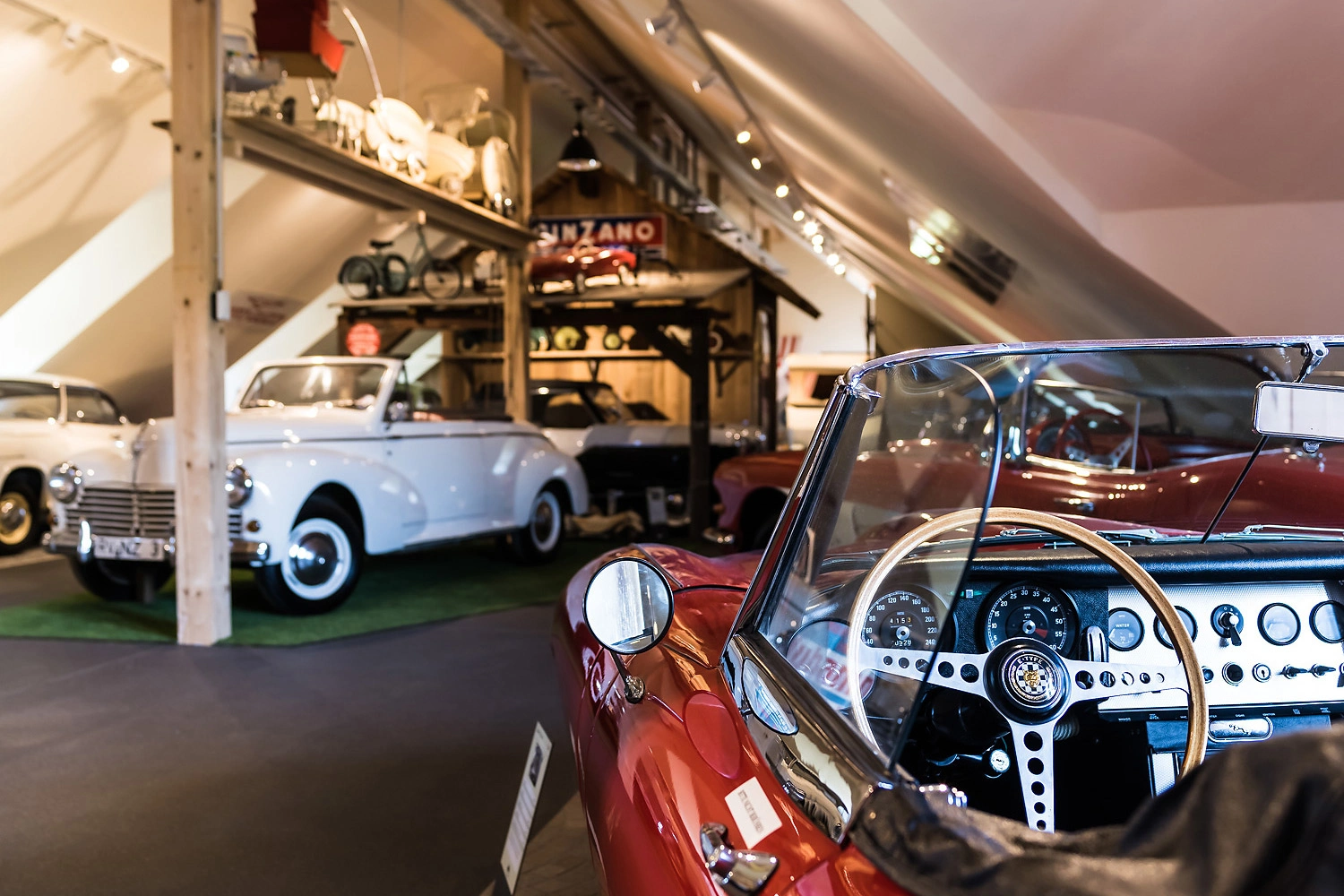 Ein roter Oldtimer steht rechts an einer Wand. Zwei weiße Oldtimer stehen auf der gegenüberliegenden Seite. 