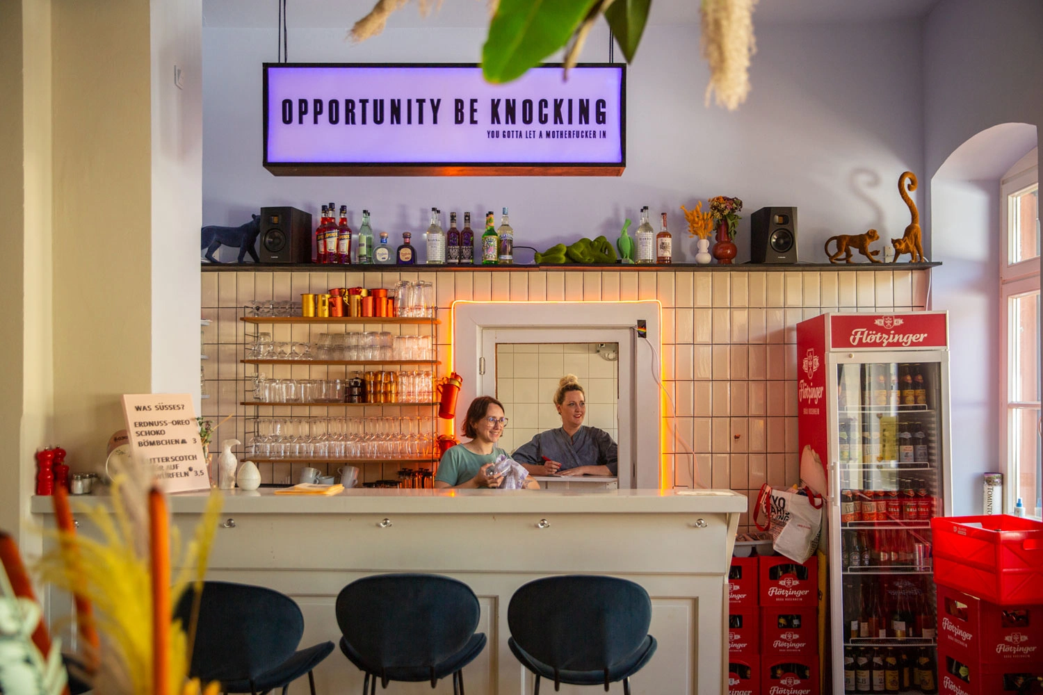 Zwei Personen stehen in einem Cafe hinter der Theke. Über ihnen hängt ein großes lila Leuchtschild. Vor der Theke stehen drei schwarze Barhocker.