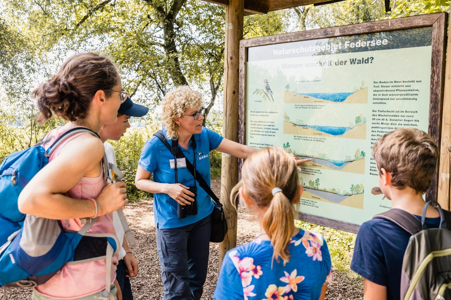 Eine Frau steht mit ihren Kindern vor einer Informationstafel in einem Wald. Eine andere Frau zeigt auf die Tafel und erklärt ihnen etwas.