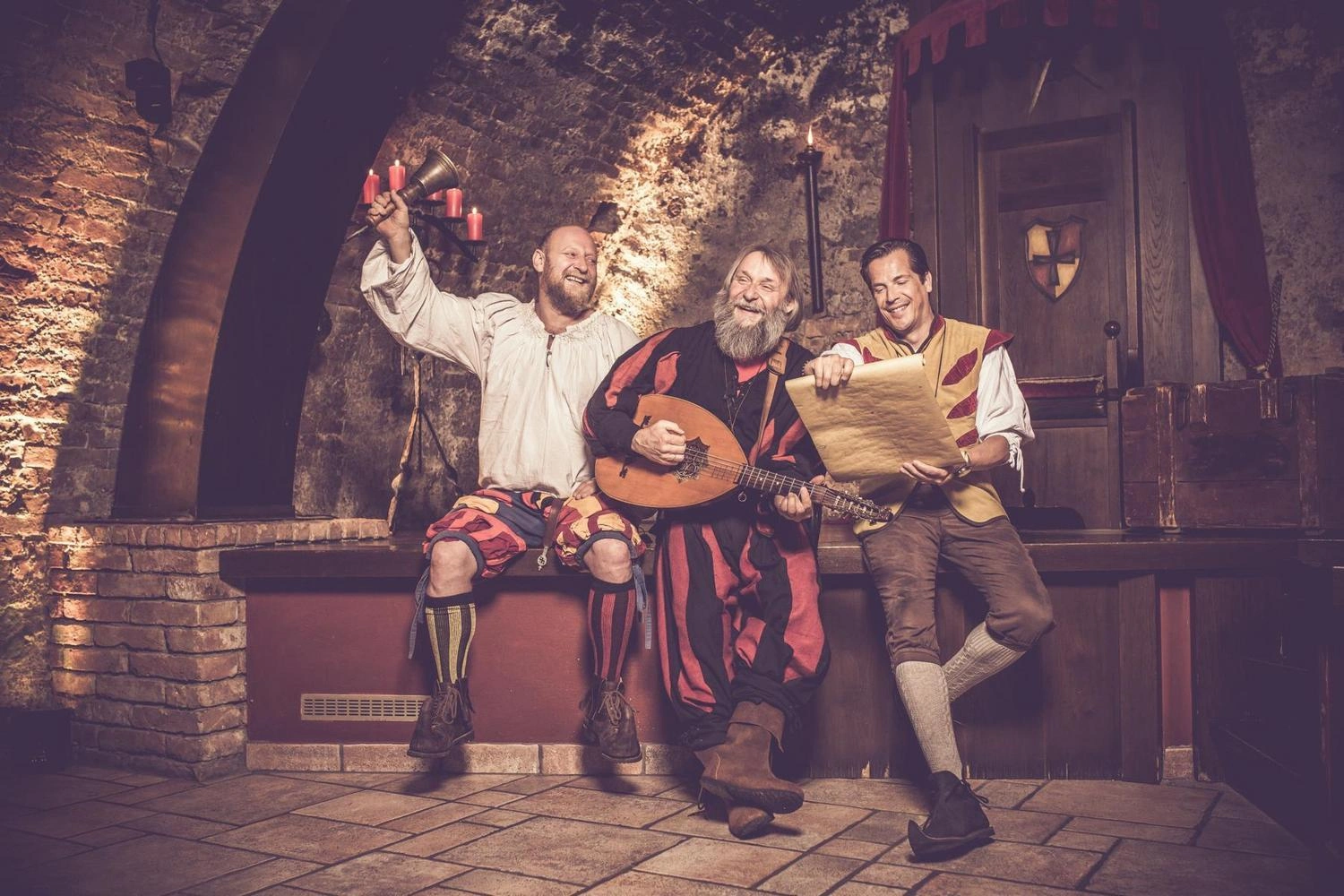 Drei Personen in mittelalterlichen Kostümen sitzen in einem historischen Raum mit Steinwänden, halten eine Laute und ein Pergament.