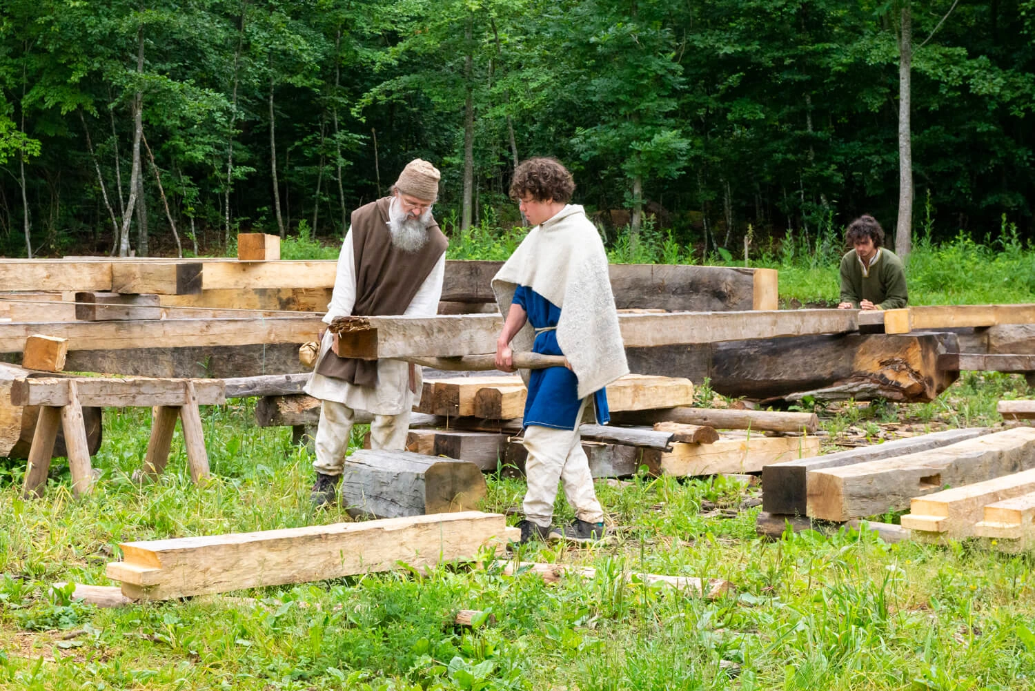 Two men in medieval clothing stand in front of a heavy wooden beam. They are trying to lift the large beam with a smaller beam.