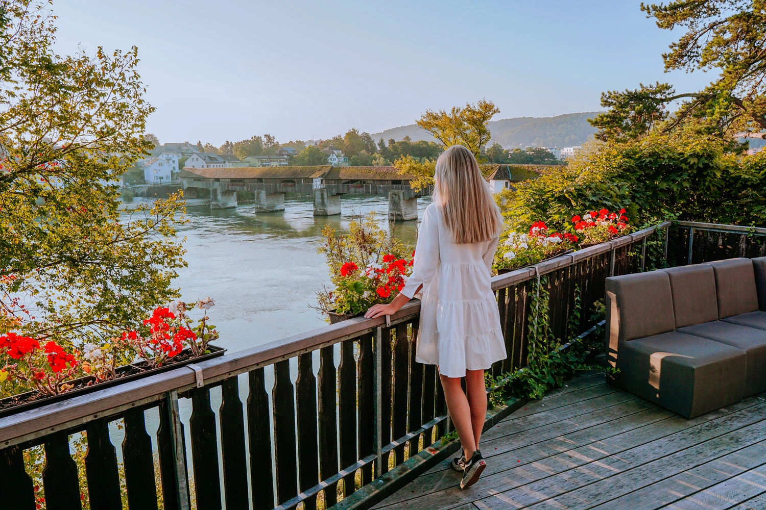 A woman stands at a railing, looking out over a river with a long covered wooden bridge.