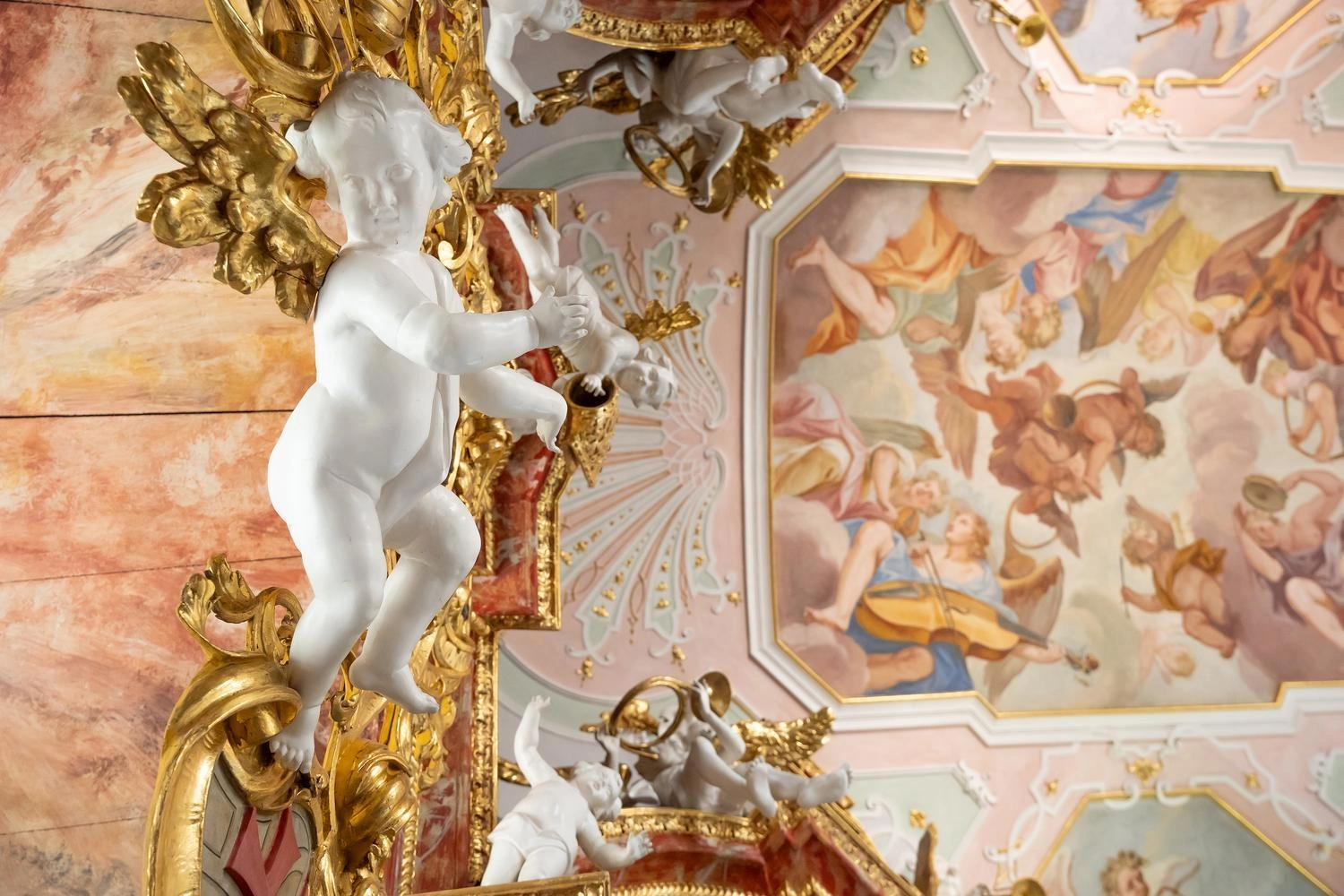 Detail einer barocken Kirchenverzierung mit weißem Engel und goldenen Ornamenten vor einer Decke mit farbigem Fresko.