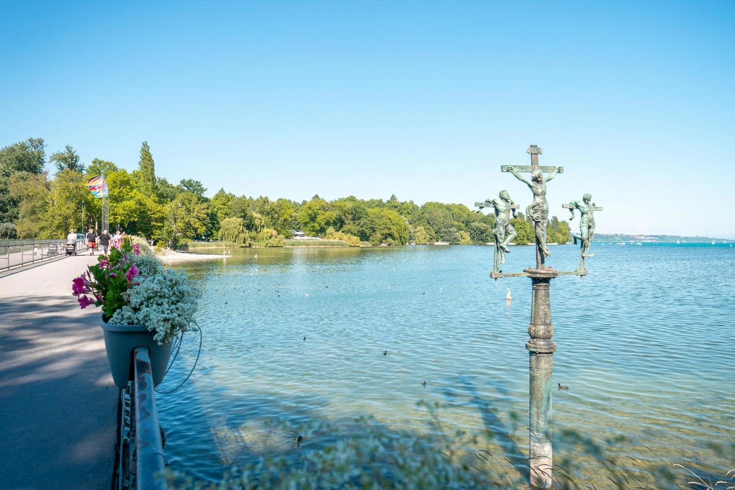 Uferpromenade mit Blumen, klarem Bodenseewasser und einer kunstvollen Kreuzigungsstatue im See, dahinter grüne Bäume und blauer Himmel.