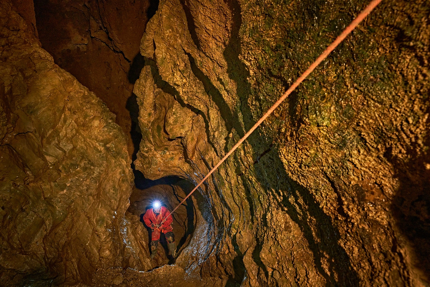 Person seilt sich mit Stirnlampe und rotem Overall an einem Seil durch eine enge, felsige Passage in der Tiefenhöhle der Schwäbischen Alb ab.