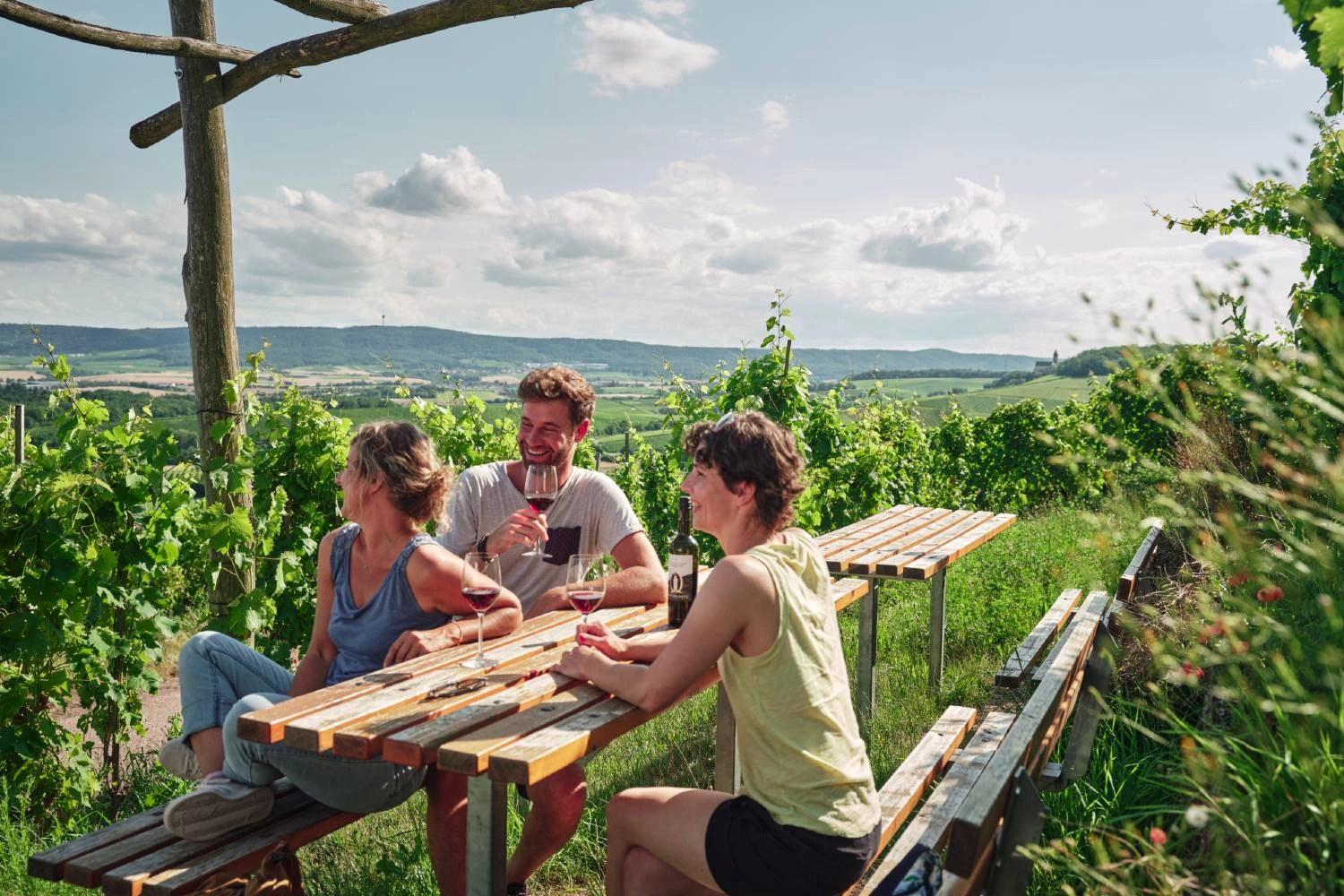 Drei Personen sitzen an einer Tischgruppe zwischen Weinreben und genießen Wein mit Blick über die Weinberge des Kraichgaus. Ein Weinausschank inmitten der Natur lädt zum Verweilen ein.