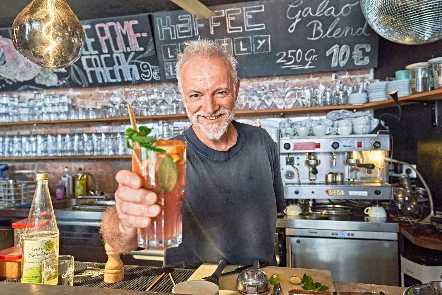 Barista hands a glass with a refreshing drink and mint over the counter in a café with a coffee machine and labeled board in the background.