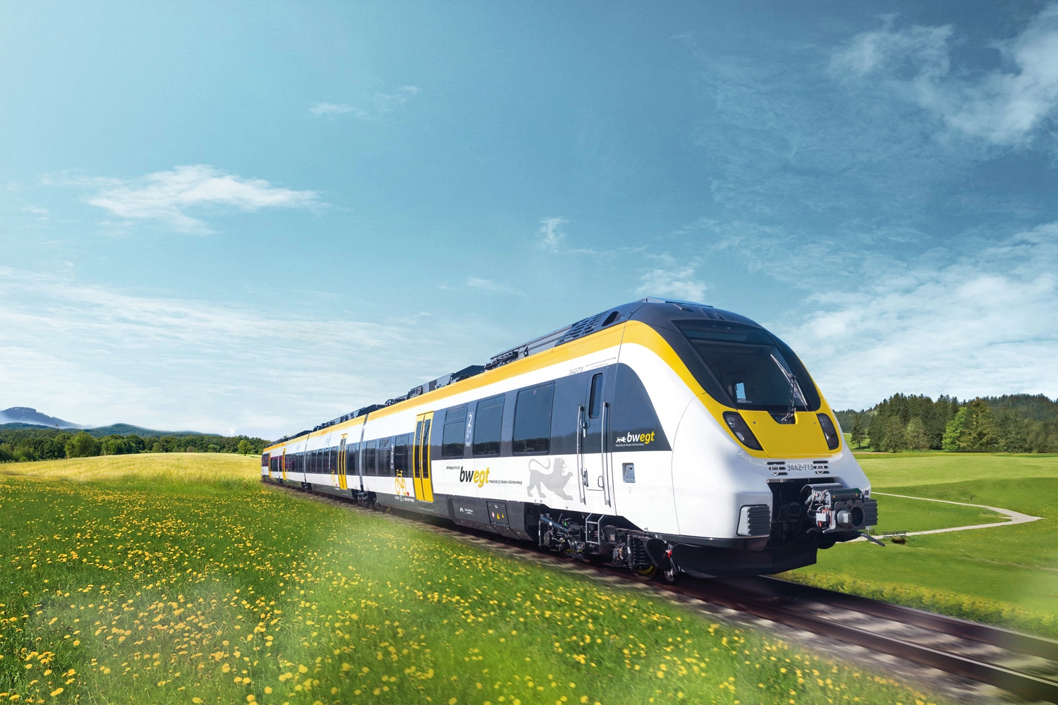 A modern white passenger train with yellow and grey accents travels through a picturesque landscape. 