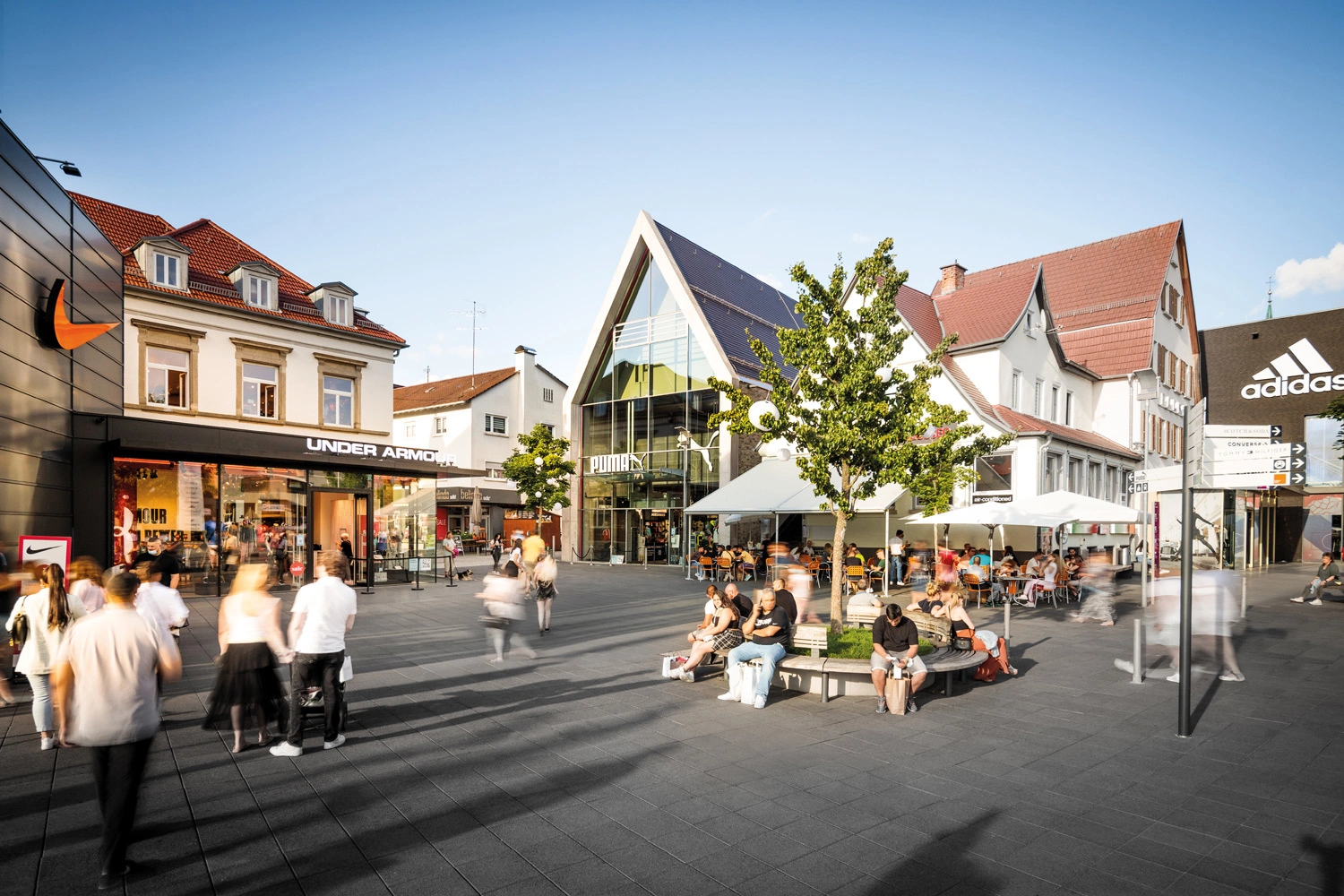 Um einen kleinen Platz herum sind verschiedene Marken-Stores platziert. Zahlreiche Menschen flanieren und shoppen da. Unter einer Linde ruhen sich manche aus.
