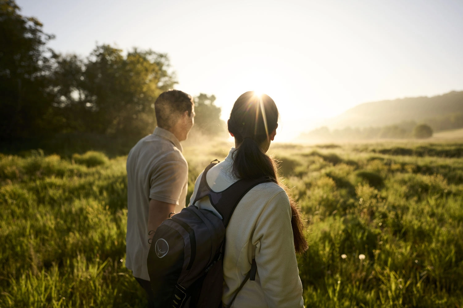 Zwei Personen mit Rucksäcken stehen auf einer Wiese und blicken in die aufgehende Sonne.