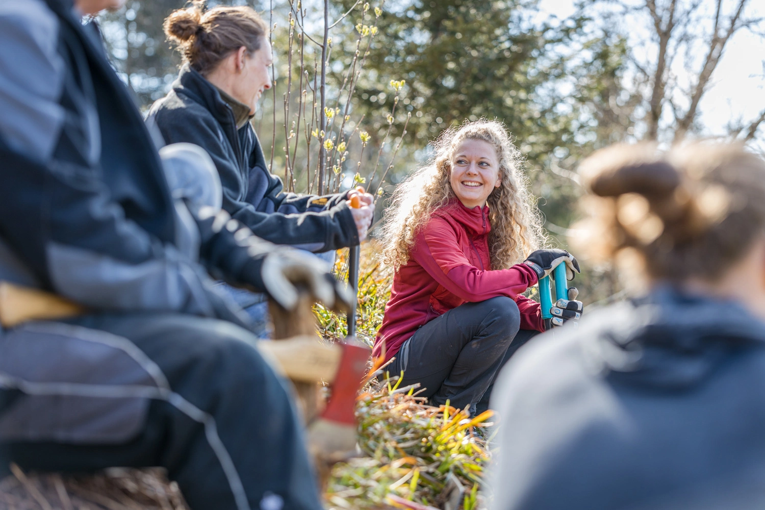 Drei Personen sind im Freien bei einer Gartenarbeit zu sehen. Die Person in der Mitte mit lockigem Haar trägt eine rote Jacke und hält ein Werkzeug. Die Umgebung ist grün und zeigt Pflanzen.