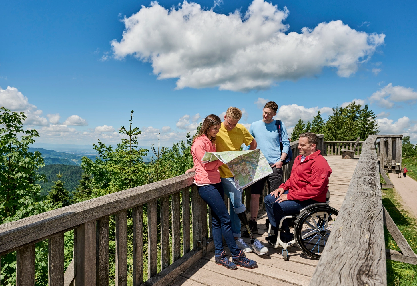 A group of hikers, one of them in a wheelchair, stands on a wooden bridge and studies a hiking map.