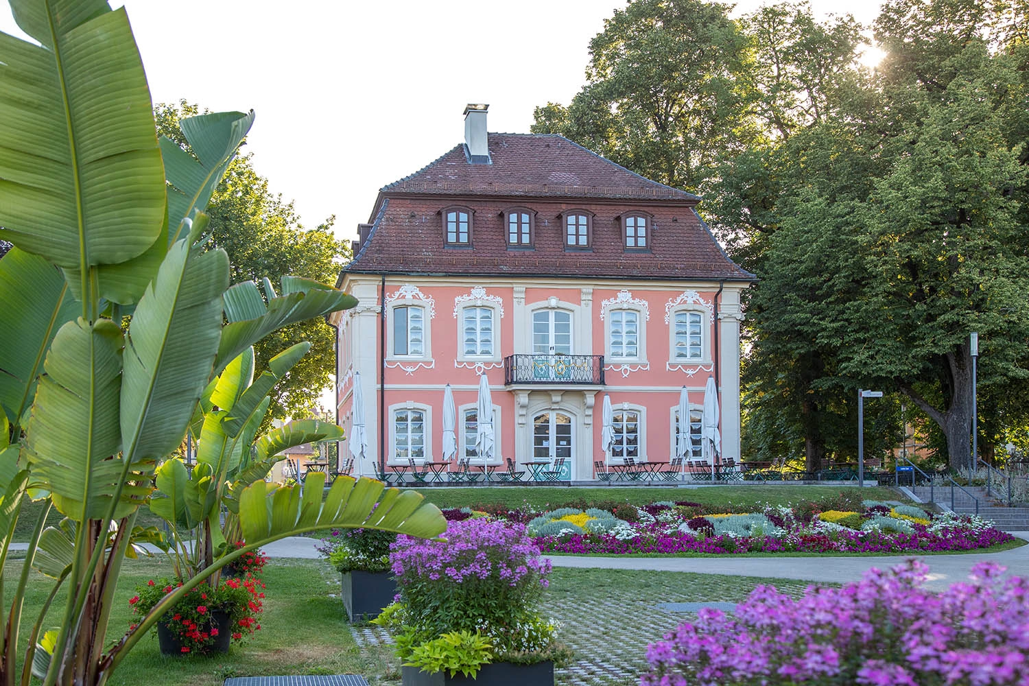 A romantic little castle with a pink façade and white windows stands in a lush garden with flowering plants and banana palms.