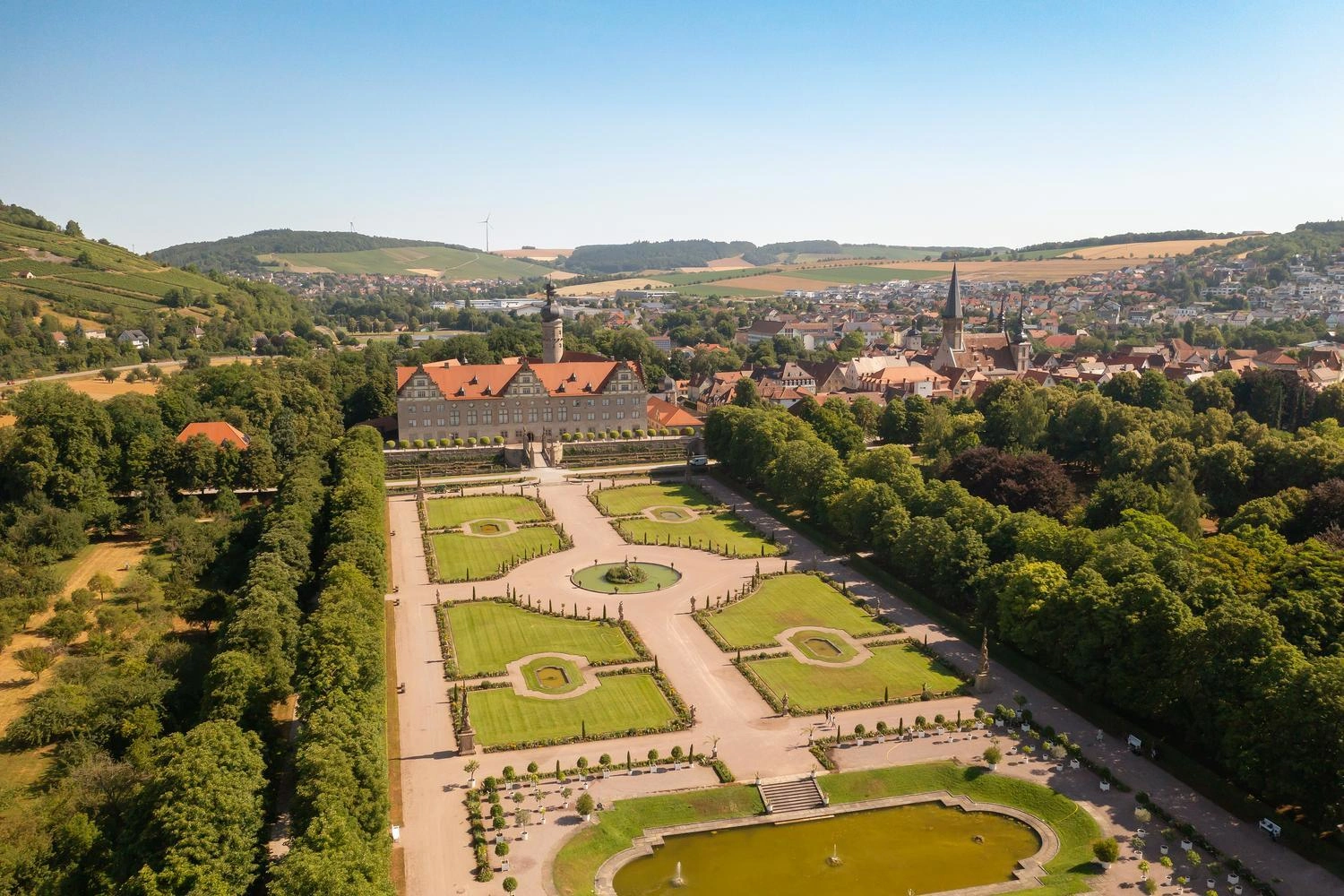 Luftaufnahme des barocken Schlossgartens Weikersheim mit symmetrischen Grünflächen, Wasserbecken und dem Schloss im Hintergrund, umgeben von Hügeln und Stadt.