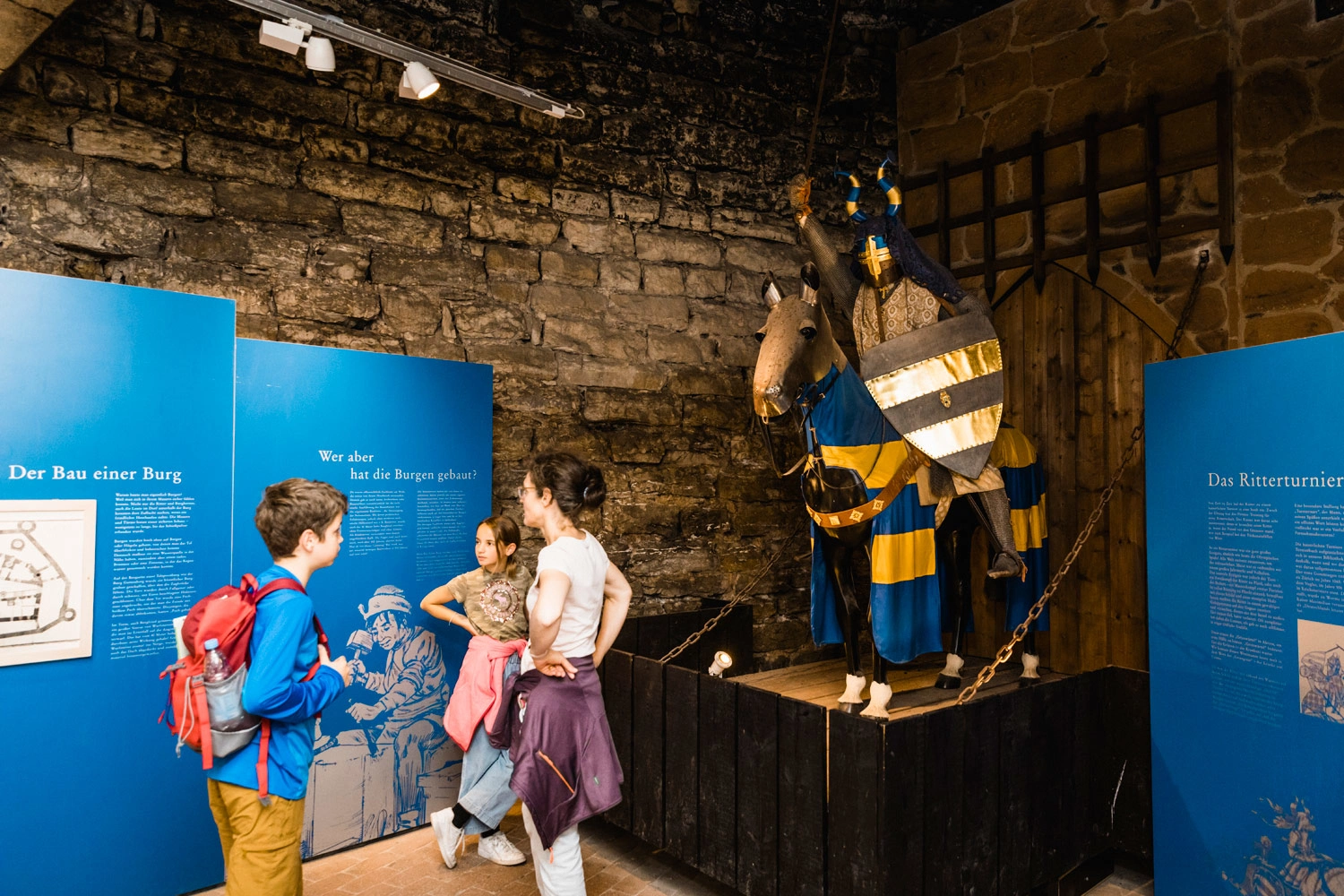 Eine Mutter mit ihrem Sohn und ihrer Tochter in einem Burgmuseum. Im hinteren Eck steht eine Pferdenachbildung mit Ritter und Rüstung.