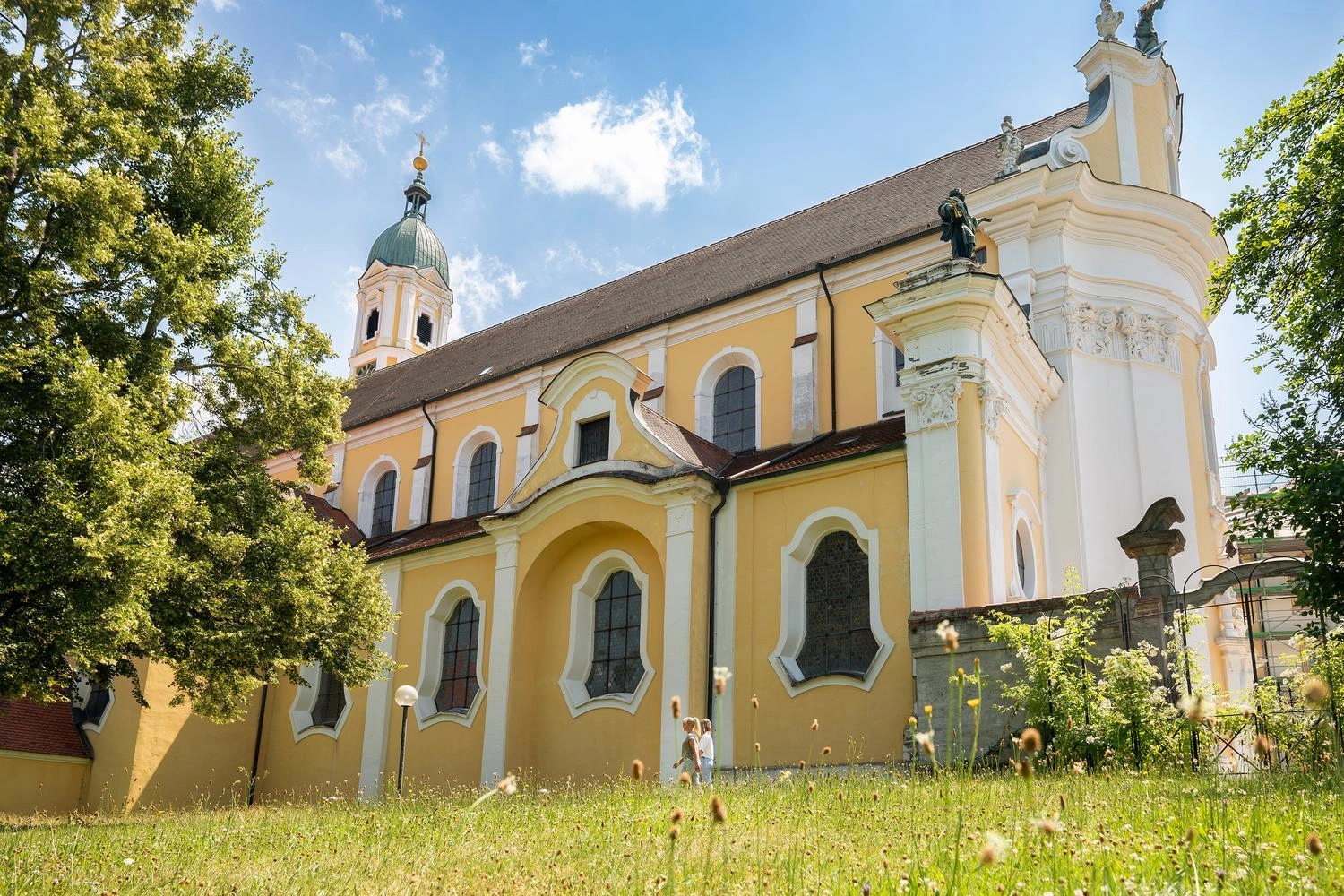 Gelbe barocke Klosterkirche mit weißen Säulen und Kuppelturm, umgeben von grüner Wiese und Bäumen unter blauem Himmel.