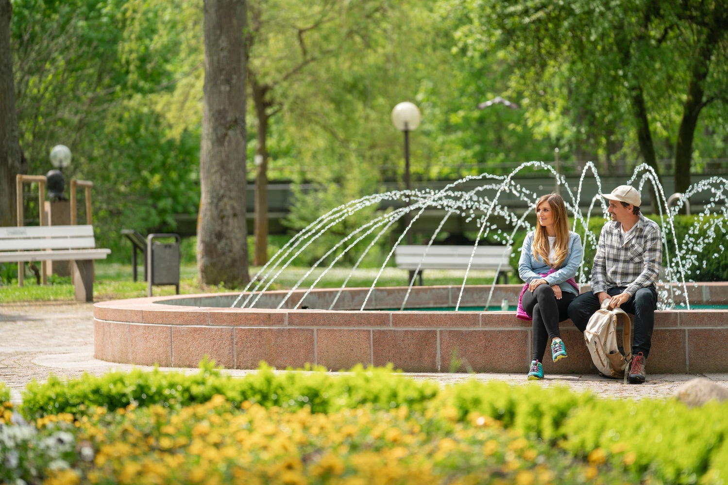 Two people are sitting relaxed at the edge of a fountain in a green park with flower beds and shady trees.