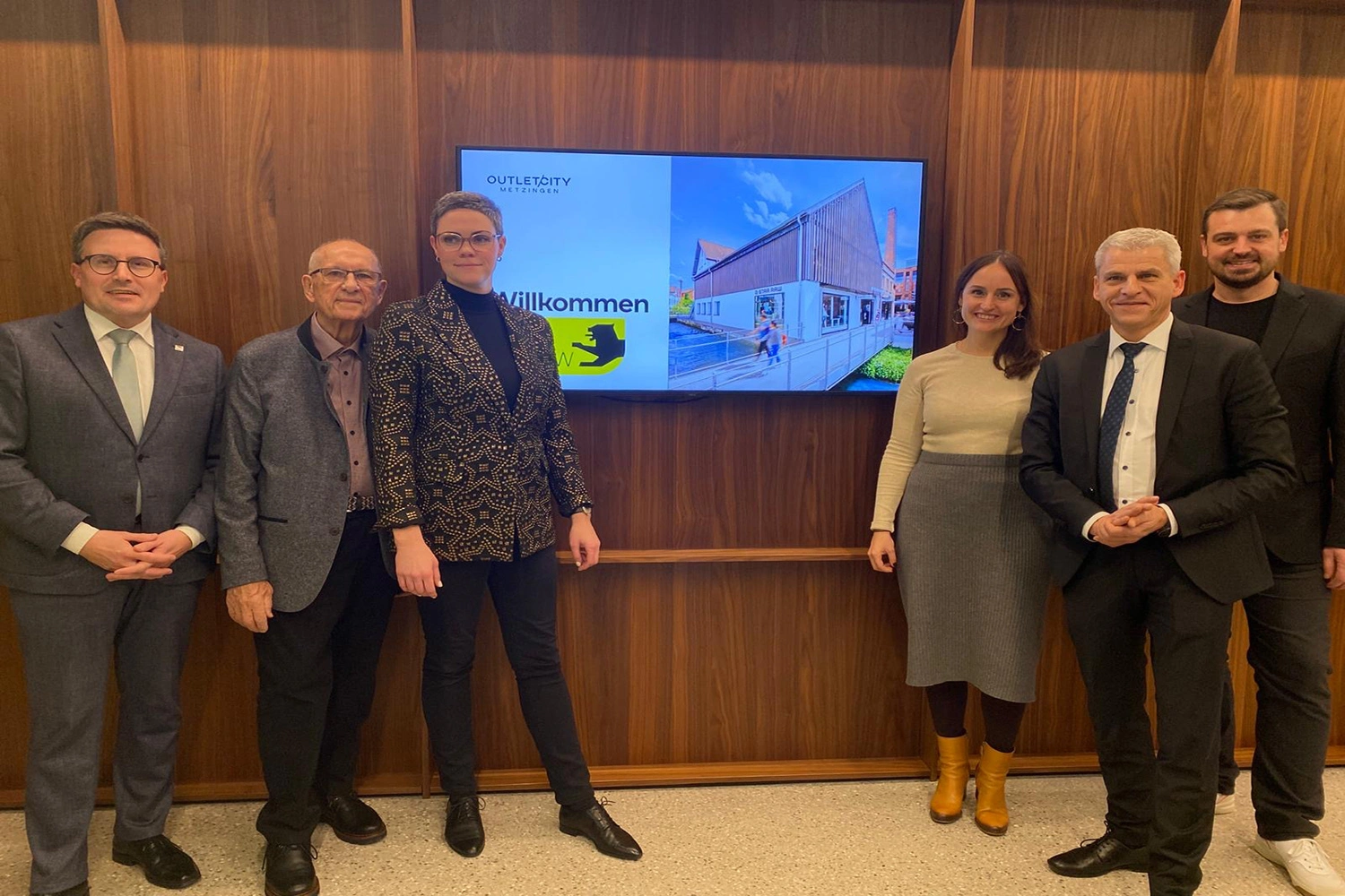 Eine Gruppe Menschen in Business-Outfit steht vor einem Bildschirm an einer Holzwand.